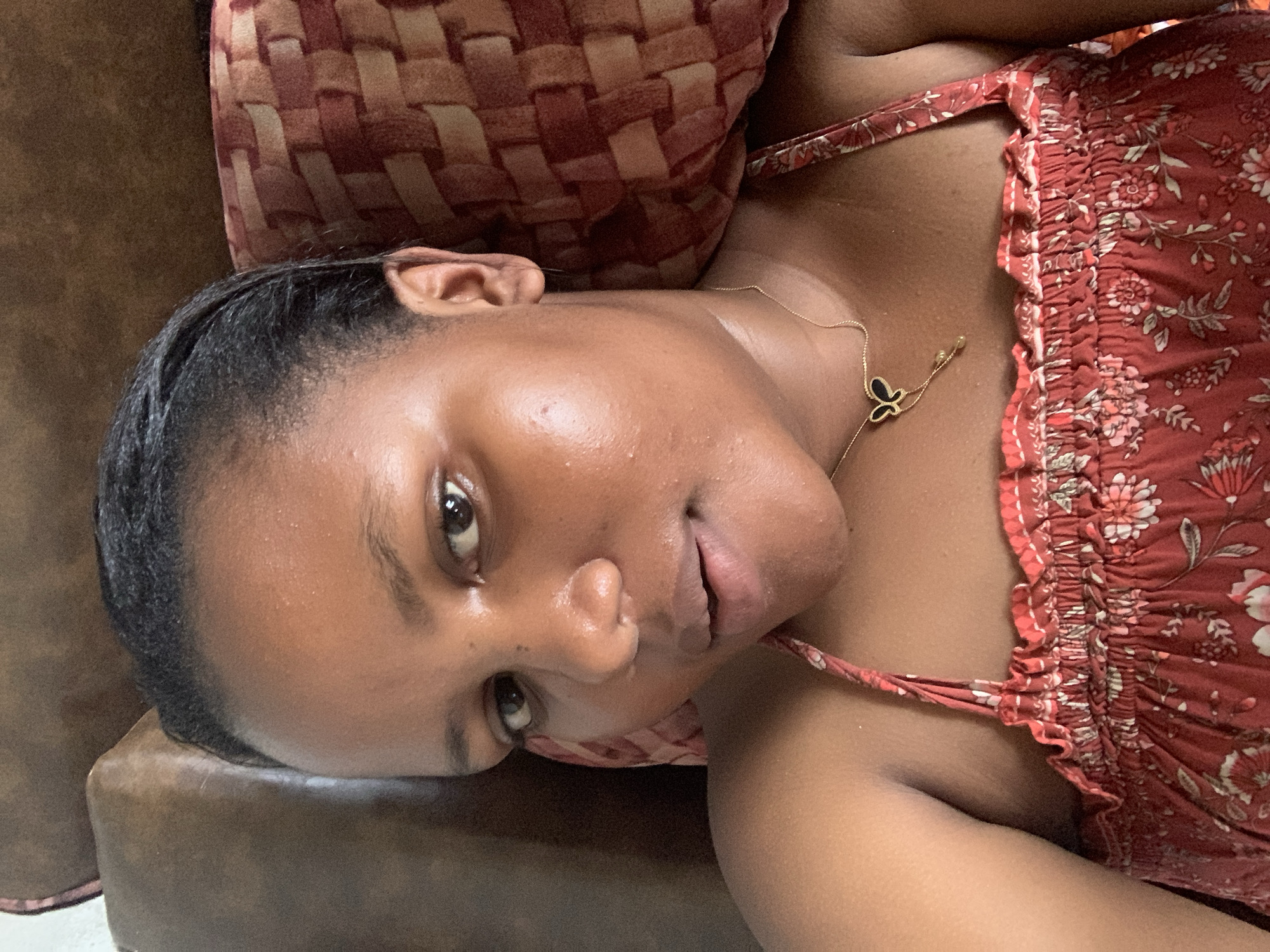 A woman lies on a brown couch, with her head resting on a red and brown patterned pillow. She is wearing a red floral dress with spaghetti straps and a gold necklace with a butterfly pendant. The location is unknown and cannot be identified as Dar es-Salaam, Tanzania from the image.