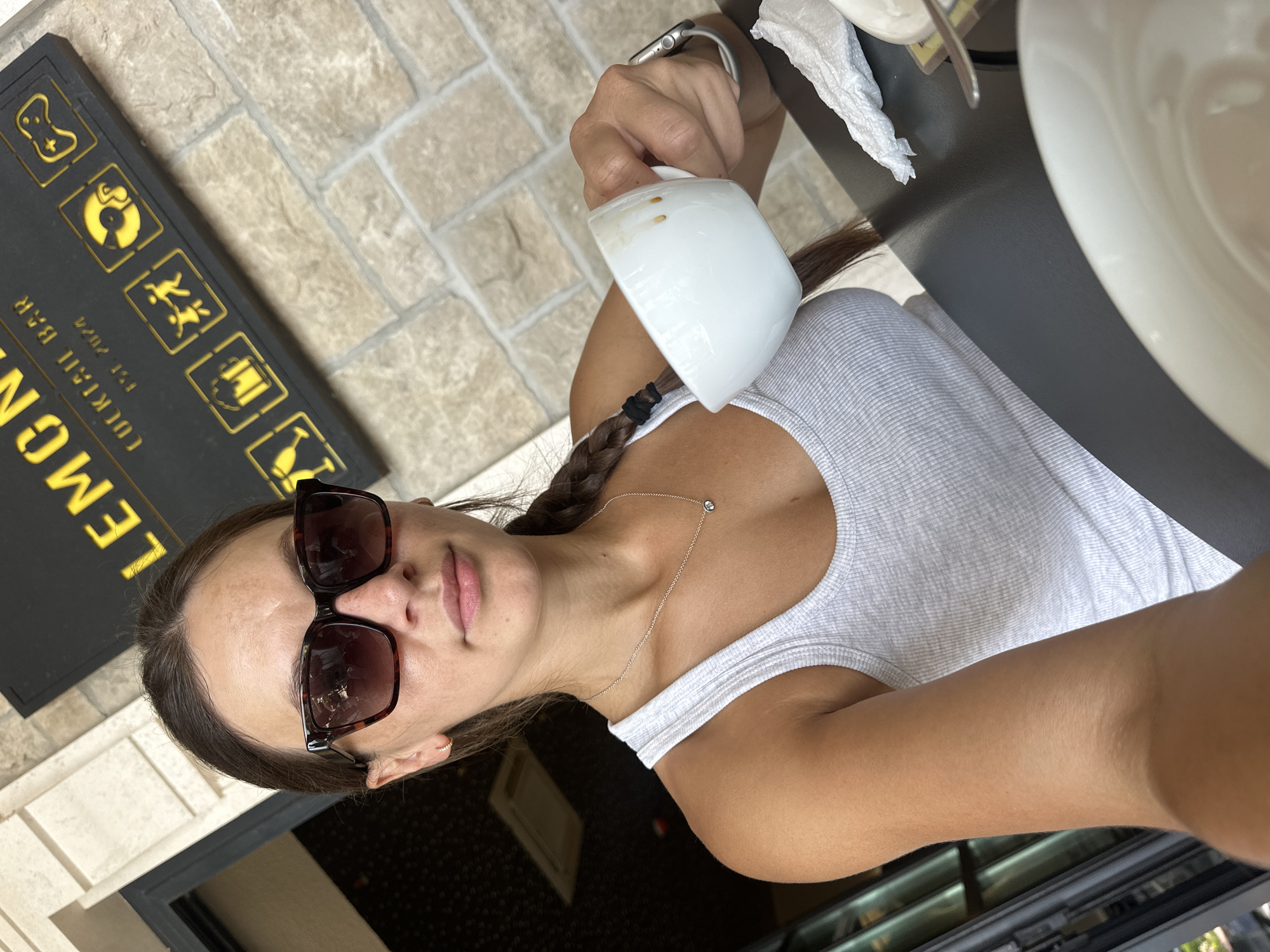 A woman with brown hair in a braid is taking a selfie at an outdoor cafe. She is wearing sunglasses, a white tank top, and a silver necklace. She is holding a white coffee cup in her right hand. The background shows a stone wall and a sign that reads "LEMON'S COCKTAIL BAR EST. 2024". The cafe appears to be in Rafailovići, Montenegro.