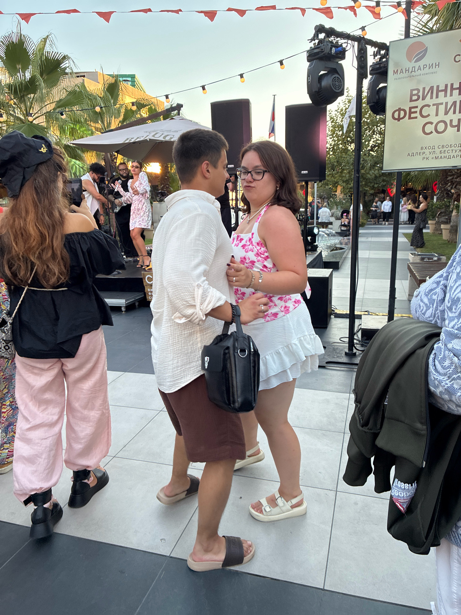 The image depicts an outdoor event in Sochi, Russia, likely at the "Mandarin" entertainment complex, as indicated by signage. In the foreground, a couple is dancing, with the man wearing a white shirt and brown shorts, and the woman in a floral print top and a white ruffled skirt. They are holding hands, and the woman is looking at the man. Behind them, on a raised stage, a band is performing, with musicians dressed in casual attire. Several people are gathered around, some observing the performance, others engaged in conversation. The environment includes palm trees, decorative lights, and a large umbrella providing shade. A banner in the background advertises a "Wine Festival in Sochi." The overall atmosphere appears festive and relaxed.