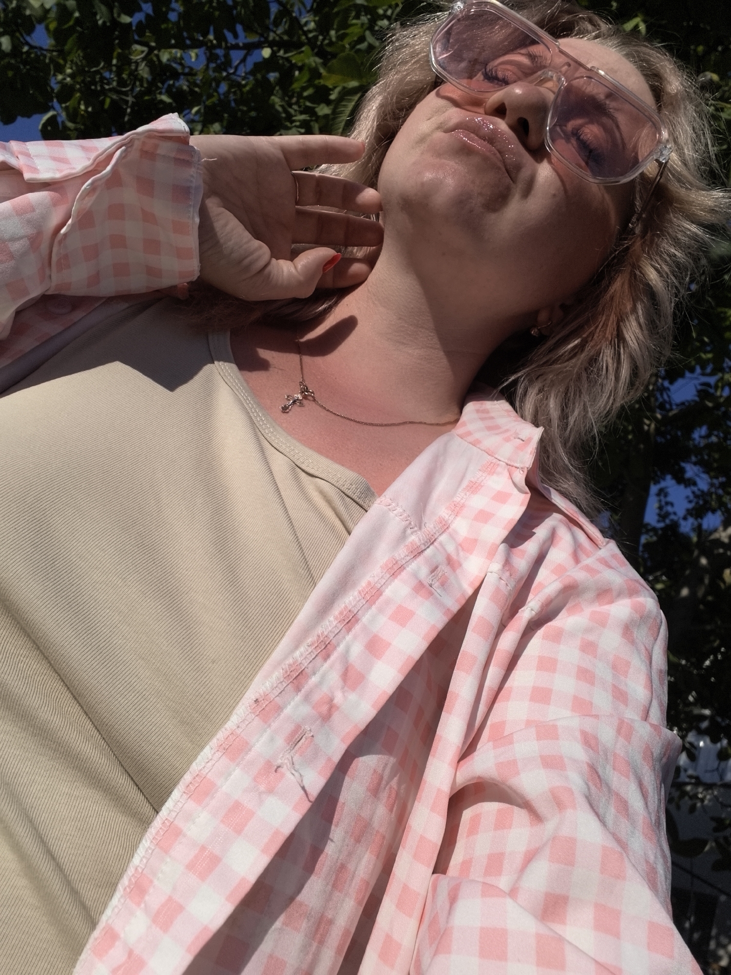 An outdoor, low-angle, close-up shot shows a woman from the chest up, wearing a beige ribbed tank top and a pink and white gingham button-up shirt. She has blonde hair and is wearing large, pink-tinted sunglasses. Her right hand is touching her chin and her left hand is blurred near her neck, possibly touching her hair. A delicate gold chain with a cross pendant rests on her chest. The background consists of dark green foliage and a bright blue sky. The lighting suggests it is a sunny day. The scene appears to be in Vityazevo, Russia, given the context provided.