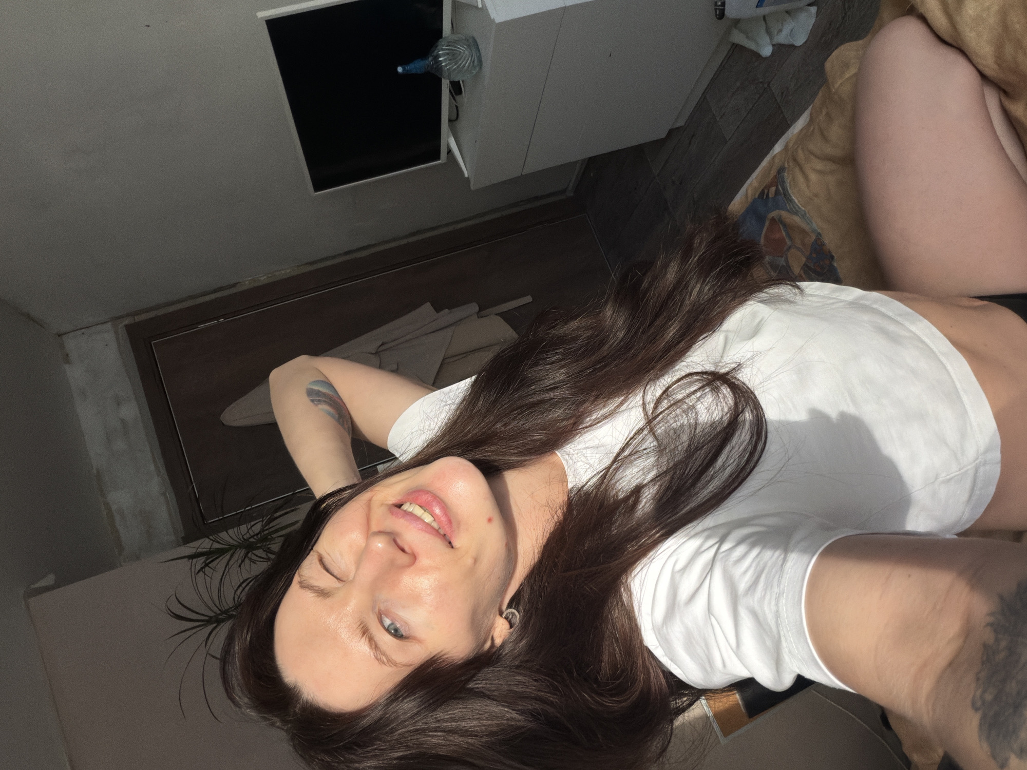 The image depicts a domestic indoor scene, identified as being located in Perm, Russia. The primary subject is an adult female, mid-shot, appearing to be taking a selfie. She has long, dark brown hair, fair skin, and blue eyes. Her left eye is closed in a wink, while her right eye is open, and she is smiling, exposing her upper teeth. She wears a white, short-sleeved crop top and dark bottoms; her bare legs are visible. Her right arm is bent with the hand behind her head, revealing a multicolored tattoo on her upper arm. A dark tattoo is also visible on her left inner forearm. A silver-colored hoop earring is present in her left earlobe.
The background features a dark wooden door and frame on the left, with a light brown garment hanging on a hook. A potted plant with green leaves is positioned behind the woman's head. On the right, a white dresser or cabinet supports a dark-screened flat-screen television. A patterned light blue glass bottle with a stopper is placed on the dresser to the right of the television. The floor is covered with dark, possibly wooden or tiled, planks. The woman is seated or reclining on a surface draped with a brown and patterned blanket. Strong natural light, consistent with sunlight, enters from the left side of the frame, illuminating the subject and portions of the room.