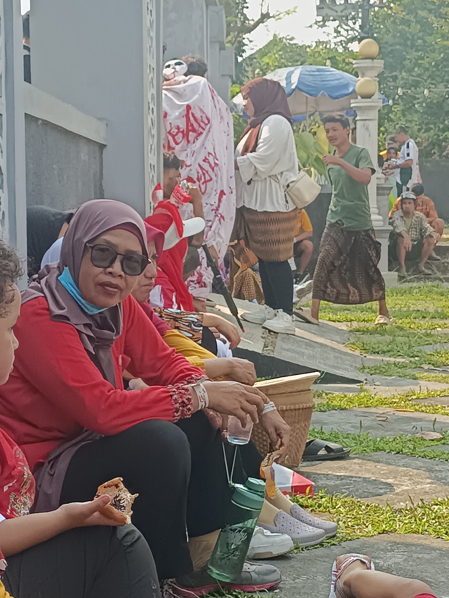 In Sleman, Indonesia, a group of people are gathered outdoors near a building with an ornate pillar and what appears to be a decorative wall. In the foreground, a woman wearing a red shirt and a purple headscarf is seated and looking at the camera. She is wearing sunglasses and a light blue face mask is hanging around her neck. To her left, a child's arm is visible, holding a piece of food. Behind the woman, other individuals are seated and standing. One person is dressed in a green shirt and patterned skirt, facing away from the camera and gesturing. Another person, wearing a white shirt and brown headscarf, is also standing. Further back, several people are seated on the ground, with a large white cloth covered in red markings and a mask in the background. The ground is paved with stones and grass. The overall atmosphere suggests a public gathering or event.