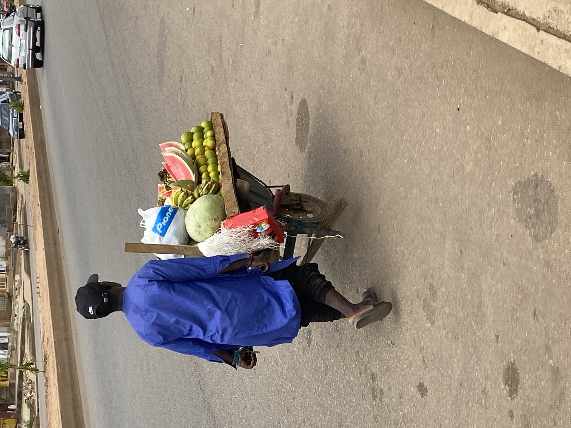An individual wearing a blue long-sleeved shirt, black pants, and flip-flops is walking along a paved road in Bukuru, Nigeria. They are pulling a cart loaded with fruits, including watermelon, bananas, limes, and a whole squash. The cart also contains a white Pioneer bag and a red plastic container. Cars are parked along the opposite side of the road, and buildings are visible in the background. The road is stained with dark spots. The individual wears a black cap.