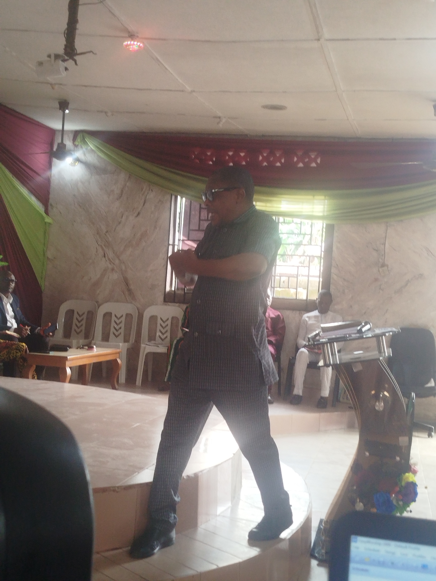 The image shows a man standing on a slightly elevated platform, possibly a stage, in a room. He is wearing a checkered shirt, pants, and dark shoes. He is facing the camera with his arms crossed or in a gesturing position. Behind him, a window is visible with decorative maroon and green fabric draped above it. A podium with floral decorations stands on the platform. Seated in the background are other individuals; one is wearing a white shirt and the other a blazer. The room has a ceiling with light fixtures, and there are chairs and a small table visible. The location is described as Unknown, Unknown.