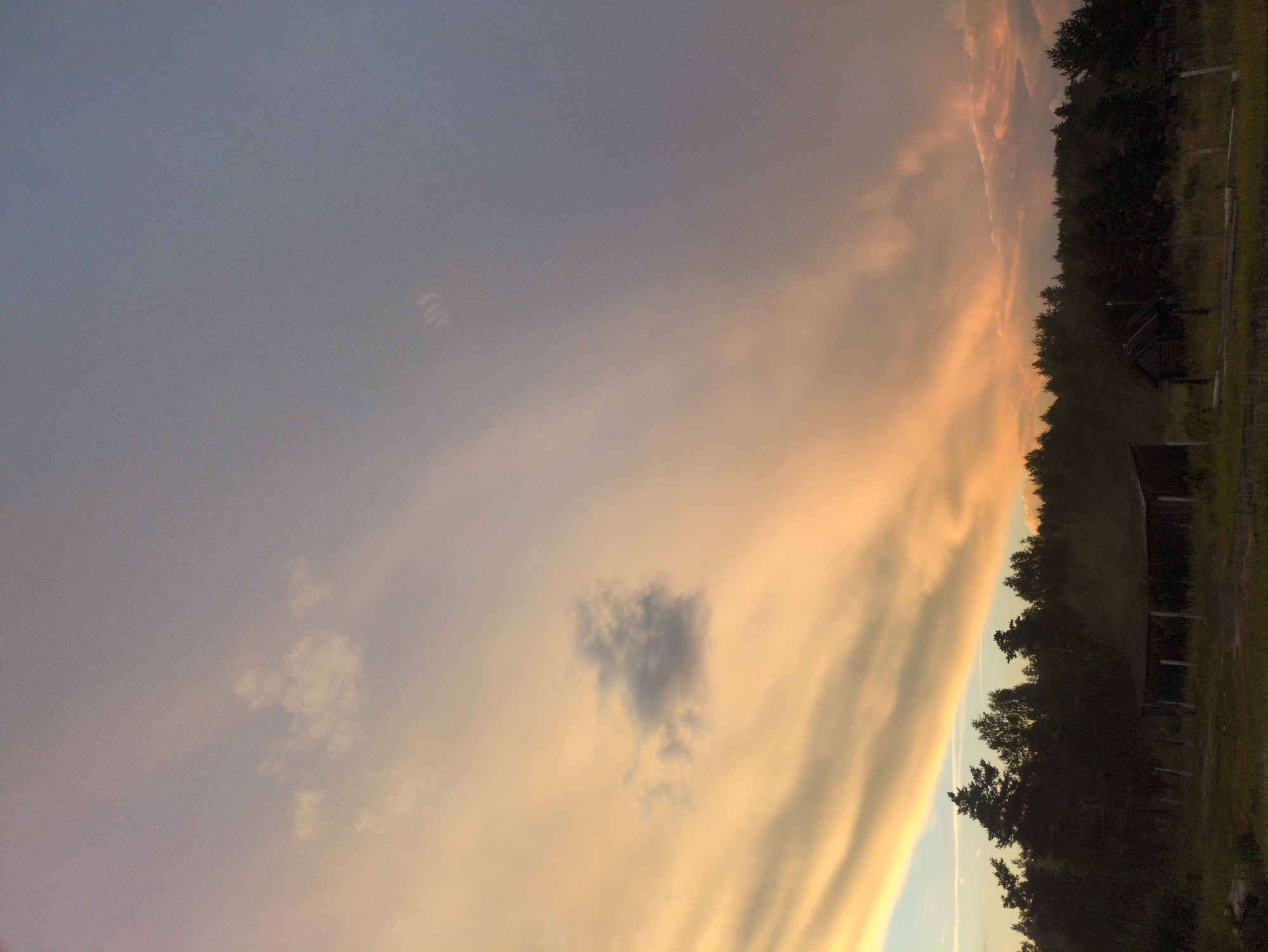 The image depicts a rural landscape under a dramatic sky during either dawn or dusk. The sky is filled with a vast expanse of clouds, painted in hues of orange, yellow, and pale blue, suggesting the sun is low on the horizon. Long, striated clouds stretch across the upper portion of the frame, with a few smaller, isolated clouds visible higher up.
In the foreground, a dense line of dark green trees forms a silhouetted forest. Beneath the treeline, a rough, open-sided wooden structure, possibly a shelter or shed, is visible. Several wooden posts are also present in front of this structure, suggesting a fenced area. The ground in front of the structure appears to be covered in dry grass and some scattered debris.
The scene is devoid of human presence. The overall atmosphere is quiet and serene, with the natural elements of the sky and forest dominating the composition. This landscape is likely located in the vicinity of Kulebaki, Russia, a region known for its natural beauty and rural character.