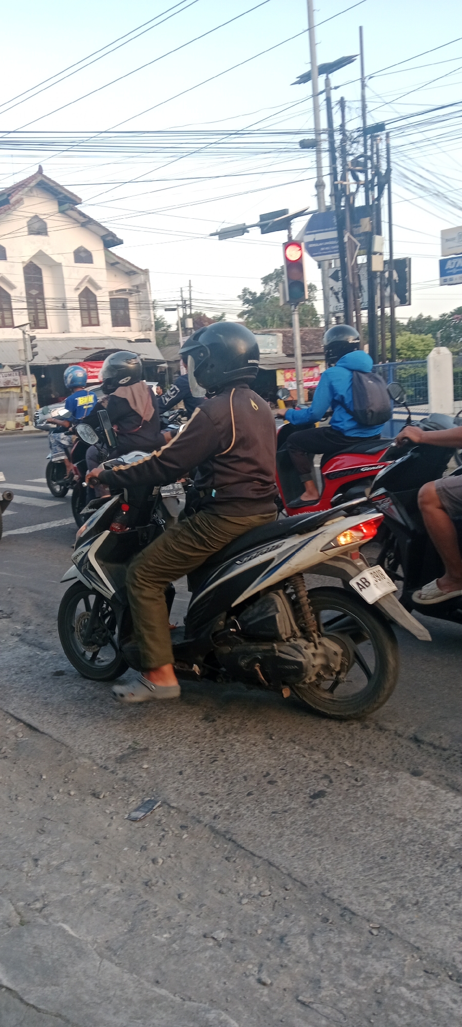 The image captures a street scene in Maguwoharjo, Indonesia, possibly at an intersection given the presence of a traffic light. In the foreground, a person wearing a black helmet and a dark jacket with yellow accents is riding a white and blue scooter. They are wearing olive green pants and grey slip-on shoes. Another person in a blue jacket and black pants, also wearing a black helmet, is riding a red scooter behind the first. A third person in a dark hijab and black helmet is on a scooter to the left, with a fourth person in a blue shirt visible further back. The road surface is asphalt with visible cracks and some debris. A white building with arched windows and a pointed roof is visible in the background, along with utility poles and a dense network of overhead wires. The sky is a pale blue, suggesting it is either early morning or late afternoon. A license plate on the white scooter reads "AB 3918".