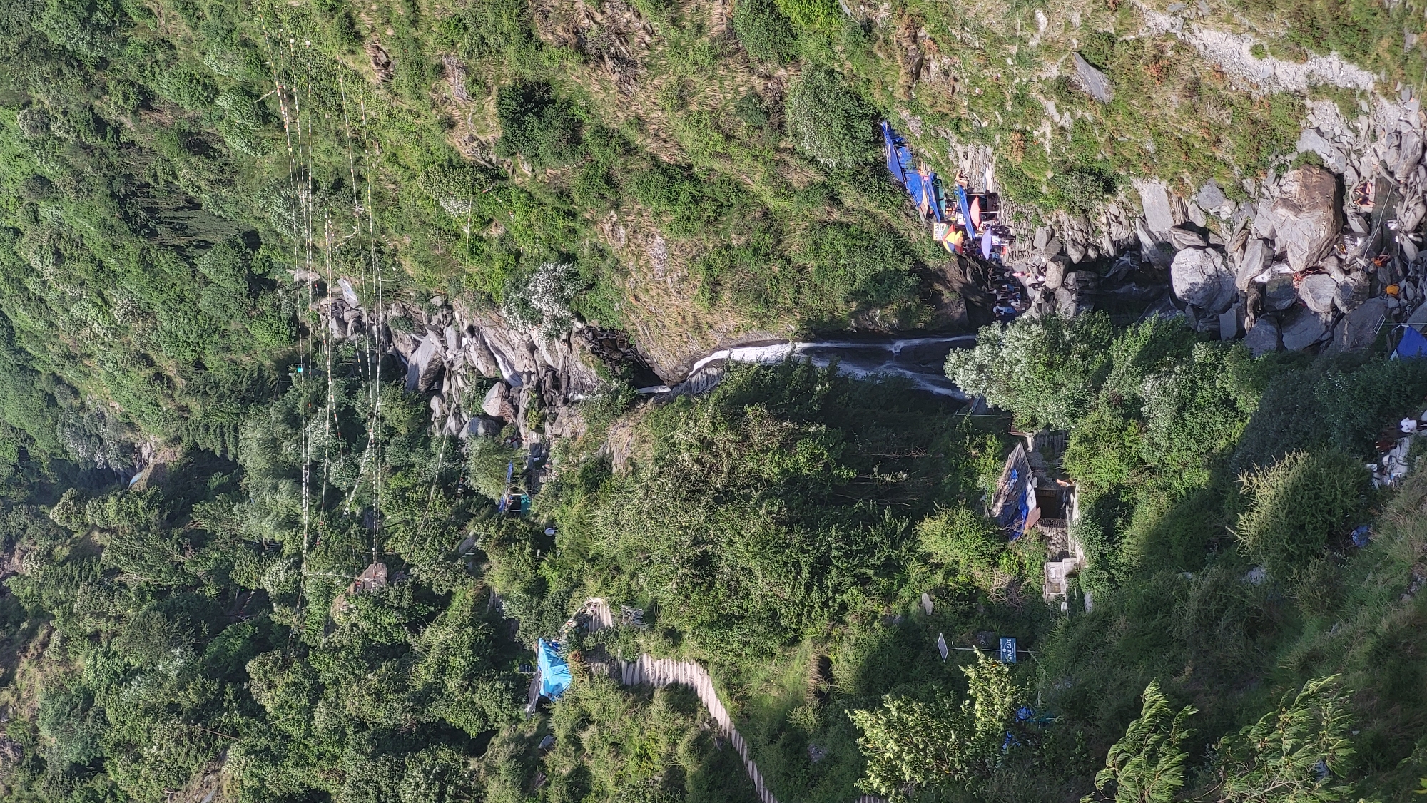 The image depicts a view of Bhagsu, India, from an elevated perspective. A stream or small river flows through a rocky channel, flanked by dense vegetation consisting of trees and shrubs. A small waterfall is visible along the stream. People are gathered near the water, and along the banks are colorful tents or stalls, suggesting a commercial or recreational area. A path, likely a series of steps, winds through the vegetation leading to structures, and perhaps a café. Power lines are also visible in the upper left of the image.