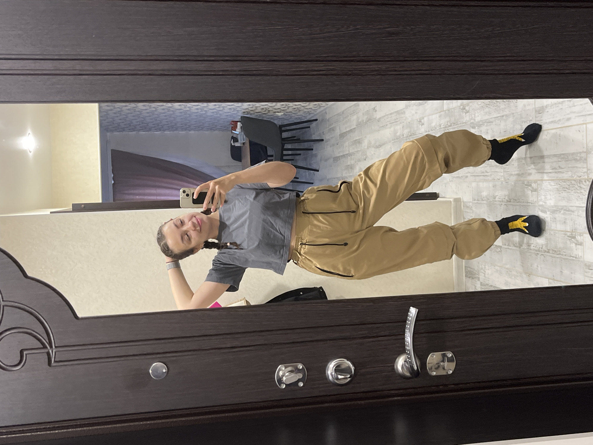 A young woman with braided hair takes a selfie in a mirror. She is wearing a gray crop top and tan cargo pants. She is standing in a hallway with a dark brown door to her left and a mirror reflecting the room behind her. The room appears to be a dining area with a table and chairs. The flooring is light-colored tile. The woman is standing with one leg bent and her hand on her head. She is holding a smartphone in her right hand.