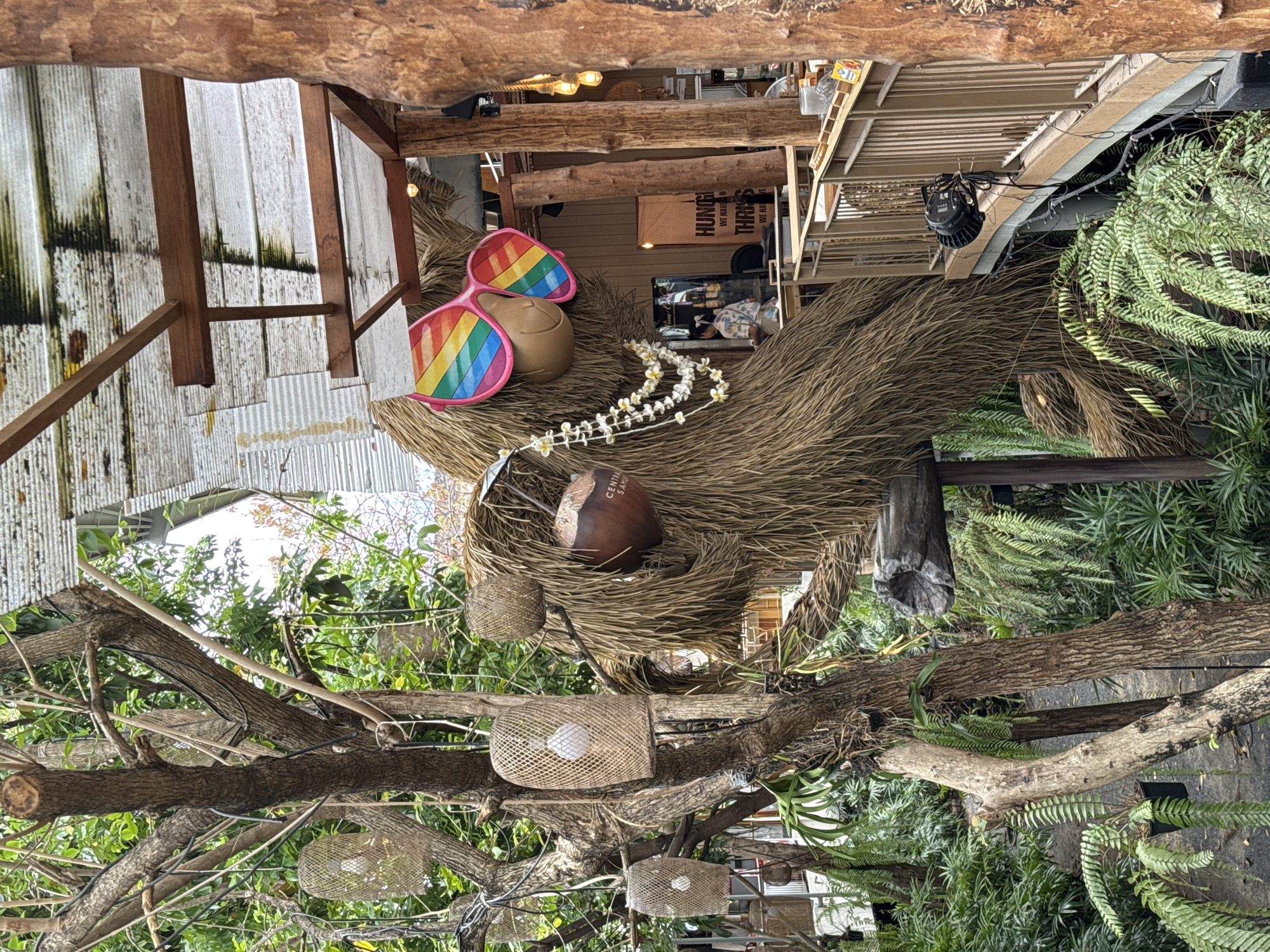 A photograph taken in Baan Chaweng, Thailand, shows an outdoor scene with natural and artificial elements. Tree branches with woven light fixtures dominate the lower half of the image. Above, a structure made of dry thatch is decorated with rainbow sunglasses, a coconut, and a lei of white flowers. The background reveals a partially visible building with signs and interior elements, including counter service. The overall impression suggests a decorated outdoor or semi-outdoor space associated with a business or event.