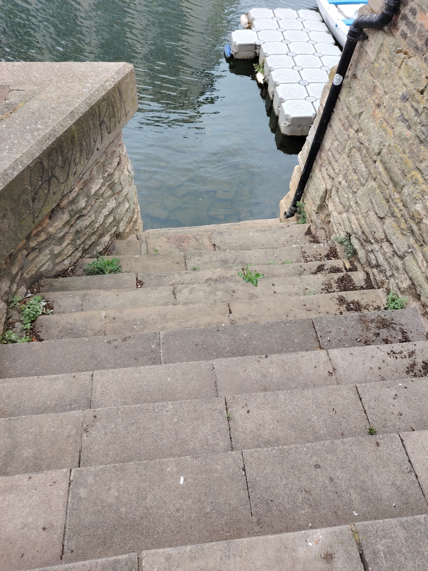 A top-down oblique view captures a flight of weathered stone steps descending towards a body of water, likely the River Nene in Peterborough, United Kingdom. The steps are constructed from large, light grey stone or concrete slabs, showing signs of wear, accumulated dirt, and sparse vegetation growing between cracks and along the edges.
On the left side, the steps are bordered by a robust concrete coping stone, beneath which is a curved, rough-hewn stone wall. Faint black graffiti is visible on the vertical face of the concrete coping, partially legible as "PETERB ALL" or "PETERBOROUGH", indicating the city's name.
On the right side, the steps are flanked by a substantial wall constructed of irregular light brown and grey stone blocks, suggesting an old embankment or bridge abutment. A black metal downpipe runs vertically along this wall, terminating near the lower visible steps. A small, circular silver sticker is affixed to the downpipe.
At the bottom of the steps, a body of calm water reflects the sky, with gentle ripples disturbing its surface. Adjacent to the right-hand wall and partially extending into the water is a modular floating pontoon system, comprised of numerous interconnected white or light grey plastic blocks. A small, white boat, possibly a rowing boat, is moored alongside this pontoon, with only its bow and a portion of its hull visible. Two white cylindrical buoys or fenders are visible near the pontoon's edge.
No people are present in the scene. The overall environment suggests an urban waterside setting, characteristic of riverfront areas in cities like Peterborough.