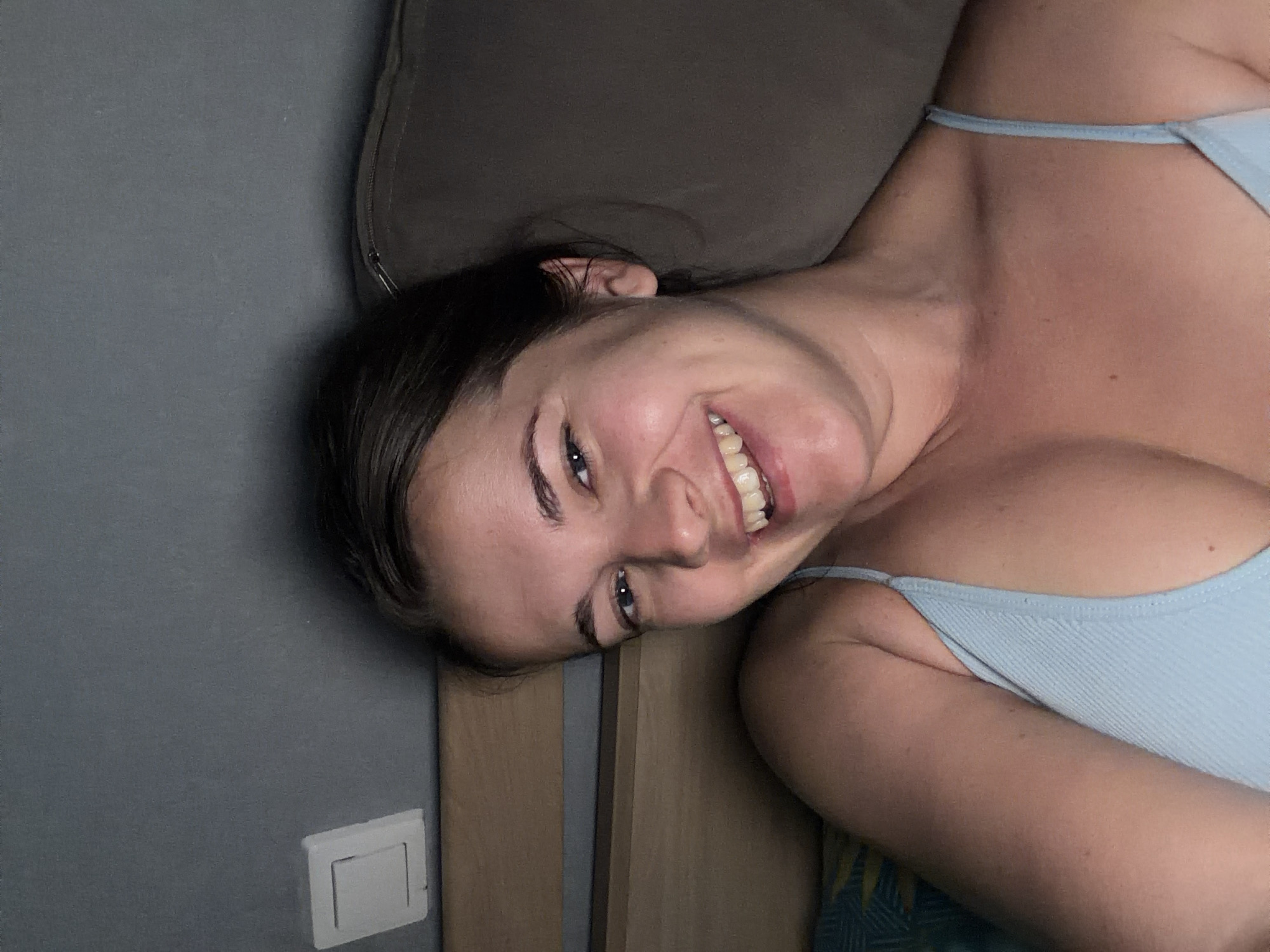 A woman with brown hair and a light blue top smiles into the camera. She is lying down, with a bed and a pillow behind her. A light switch is visible on the wall to her left. The scene appears to be indoors, possibly in a bedroom.