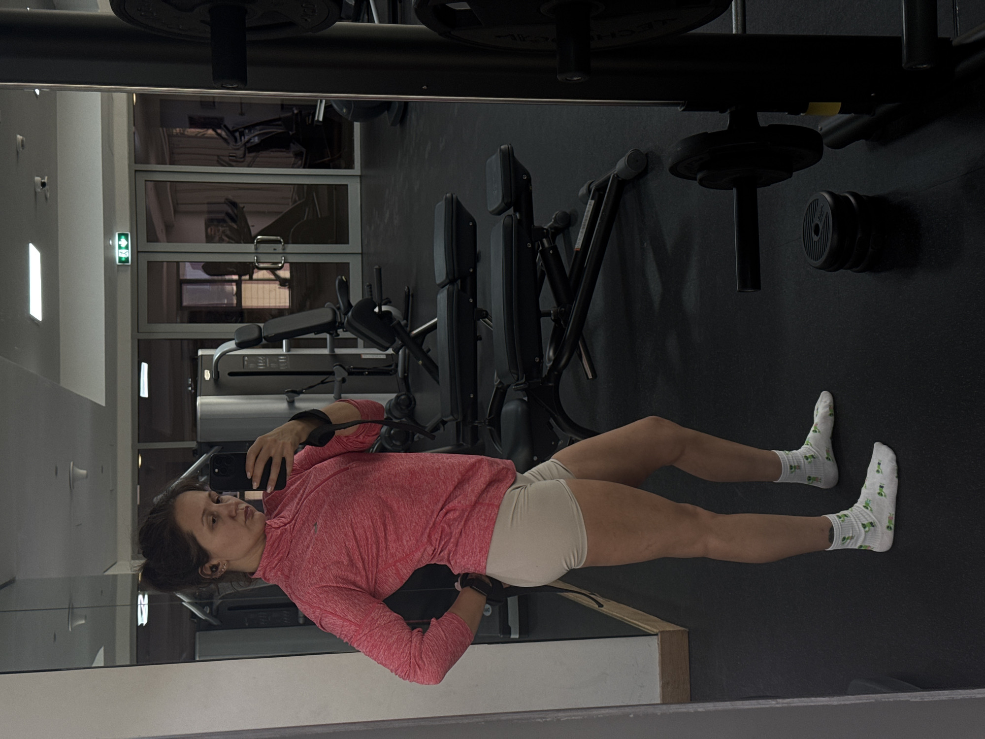 A person wearing a pink long-sleeved shirt, beige shorts, and white socks with green motifs is taking a selfie in a gym. The gym features various exercise machines and weightlifting equipment. The scene is observed through a mirror. The precise location within Dubai, United Arab Emirates, cannot be determined from the image.