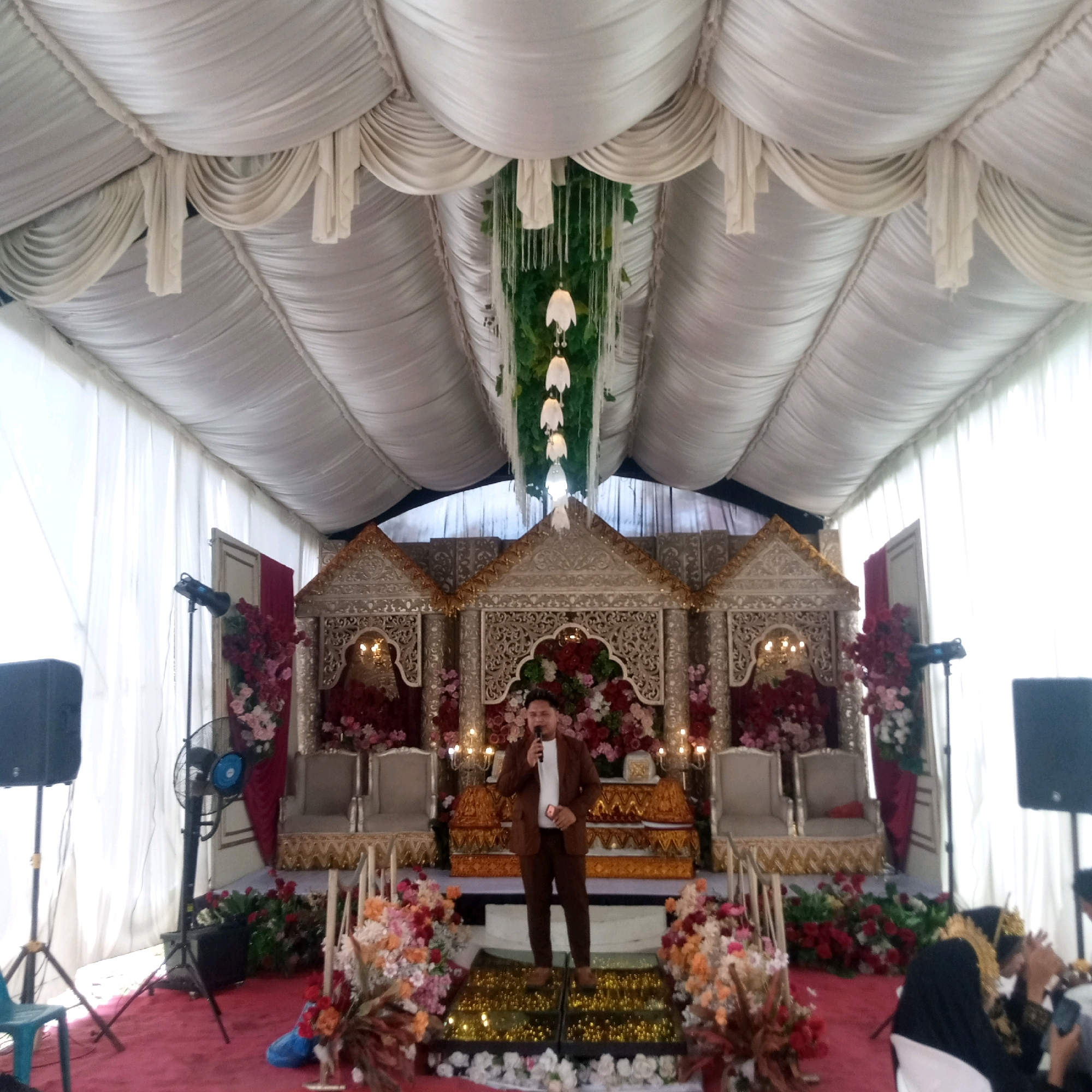 The image depicts an indoor event, likely a wedding reception, set up under a large white tent. A man in a brown suit and white shirt is standing on a raised platform, holding a microphone and speaking into it. He is the central subject of the scene. Behind him, a decorated stage area features ornate golden structures, floral arrangements of red and pink roses, and two upholstered chairs. To the left and right of the stage are speakers on stands and a standing fan. White drapes and decorative elements hang from the tent ceiling, with some greenery and pendant lights visible. The floor is covered in a red carpet, with additional floral arrangements at the front. In the background on the right, several people are seated, one of whom is wearing a traditional headpiece, suggesting a cultural celebration. The overall ambiance appears festive and formal. The location is specified as Saree, Indonesia.