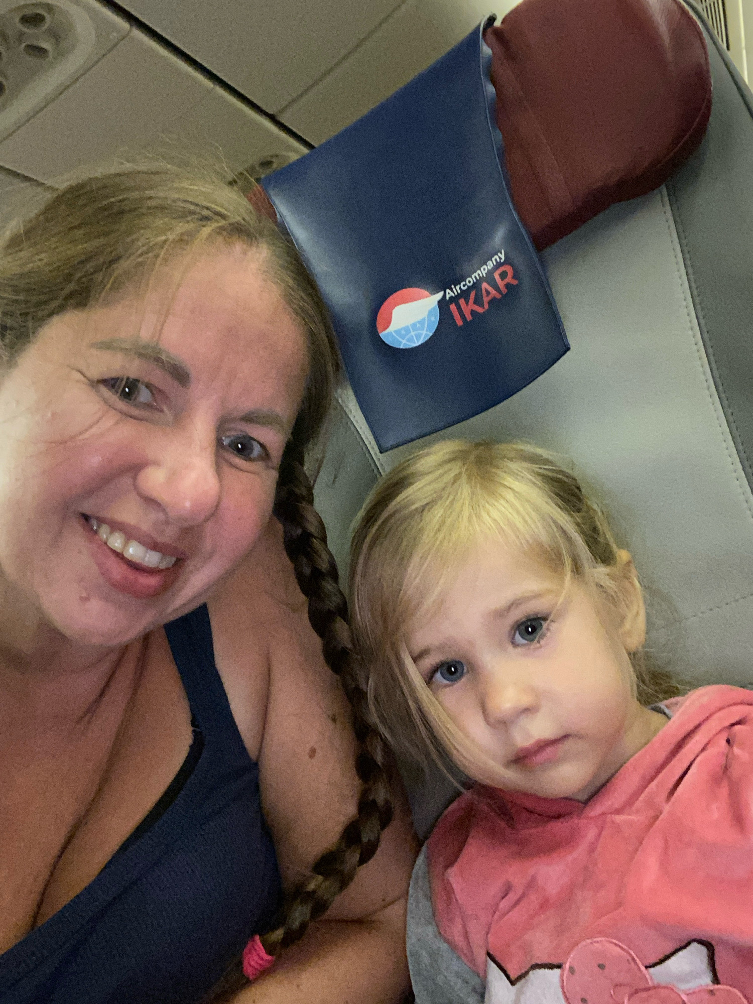 A woman and a young girl are taking a selfie on an airplane. The woman is in the foreground, smiling and looking at the camera. She has blonde hair in a braid and is wearing a blue top. The girl is to her right, looking slightly off-camera. She has blonde hair and is wearing a pink and gray hoodie. Behind them, an airplane seat with a red headrest and a blue pouch with the "Aircompany IKAR" logo are visible. The lighting suggests it is daytime. The presence of an airplane suggests this image was taken during a flight, potentially departing from or arriving at Cam Ranh International Airport (CXR) in Cam Ranh, Vietnam, which serves the city of Nha Trang and surrounding areas.