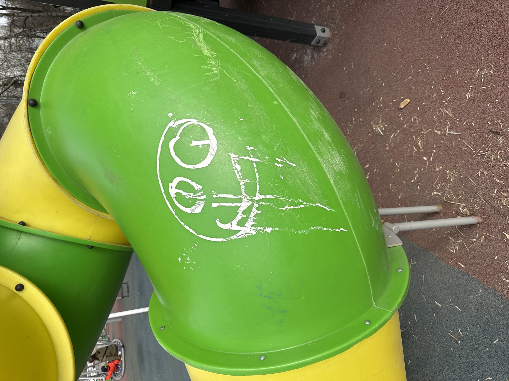 A medium shot captures a section of a green and yellow playground slide, presumably in Moscow, Russia. The slide is made of plastic and shows signs of wear and tear, including scratches and white scribbled markings on the green section. The ground around the slide is a mix of brown mulch and grey rubber surfacing. In the background, there are hints of other playground structures and trees.
