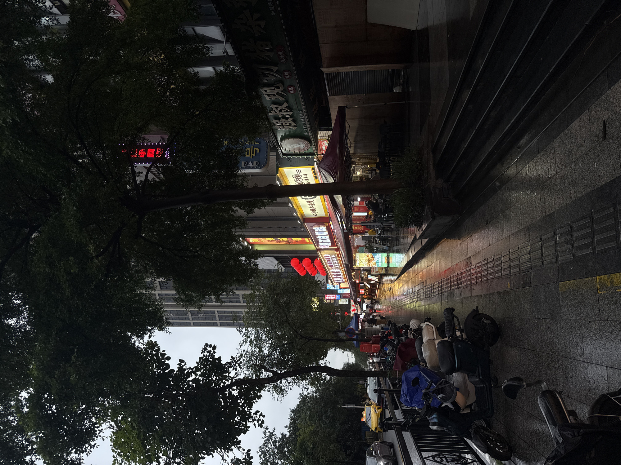 A street scene in Haizhu District, China, after rain, with wet pavement reflecting the lights of shops and streetlights. On the left side of the street, cars are driving, and a yellow taxi is visible. A row of parked motorcycles and scooters lines the sidewalk. The sidewalk is paved with textured tiles and has a tactile warning strip. On the right side of the street, storefronts display signs, some illuminated with red lanterns. People can be seen walking under umbrellas or sitting at outdoor tables in front of restaurants. The overall impression is a vibrant urban environment, perhaps in the evening or on a cloudy day, with the dampness adding a reflective quality to the scene. A tall building is visible in the background.