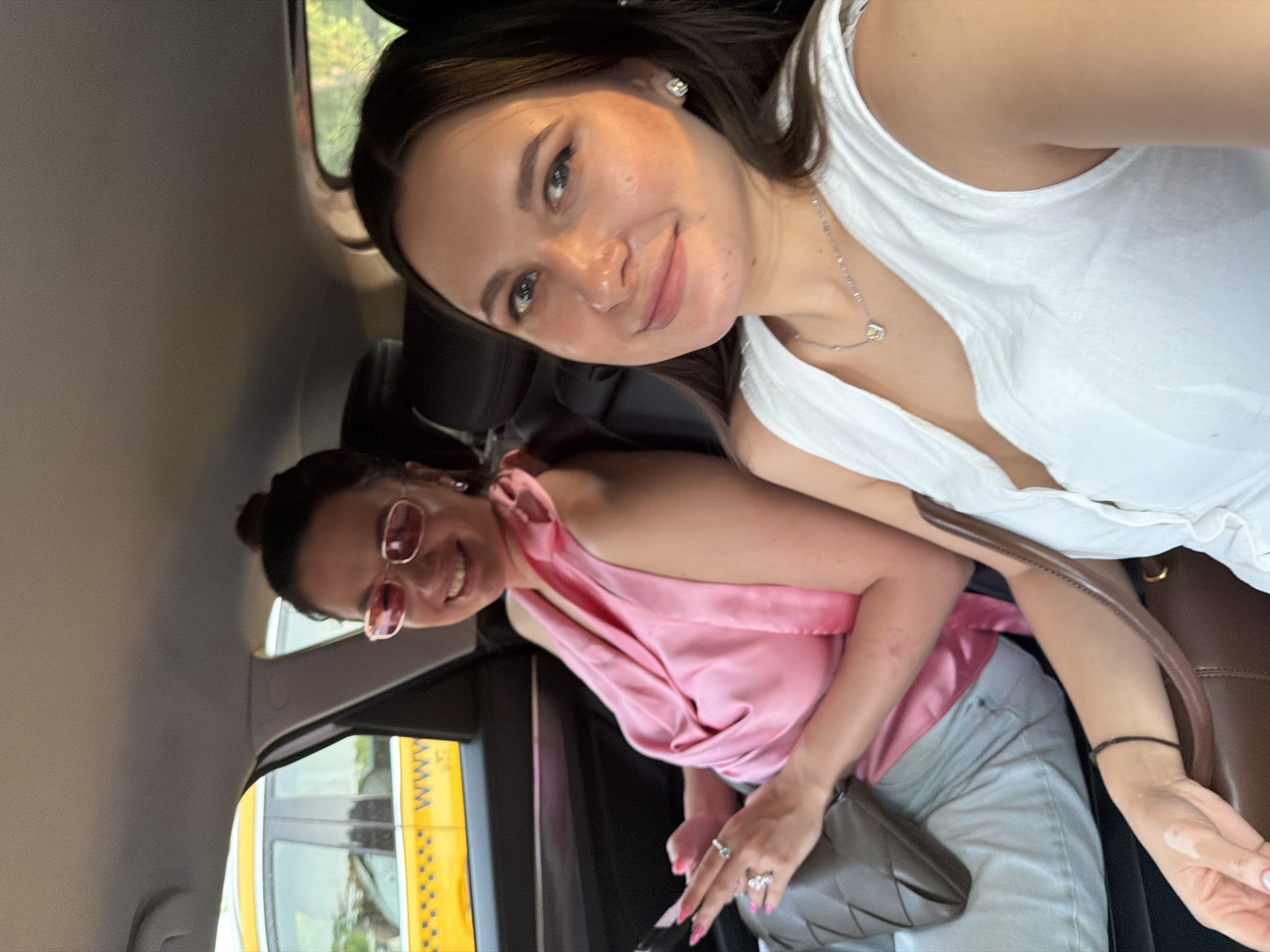 Two women are sitting in the back seat of a taxi. The woman on the left is wearing a pink top and sunglasses, and she is holding a phone. The woman on the right is wearing a white top and a necklace. The taxi is yellow and has "WWW" written on its side. The image appears to have been taken in the city of Odintsovo, Russia.