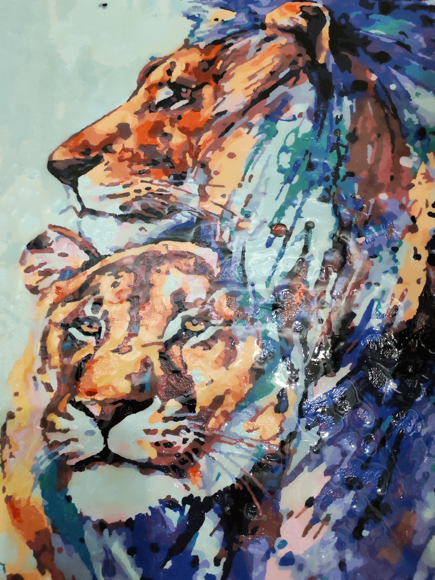 The image is a painting of two lions, a male and a female, likely a lion and lioness pair. The male lion is positioned above and slightly behind the lioness, suggesting a protective or dominant posture. The painting style is somewhat abstract or impressionistic, using blocks of color and distinct brushstrokes to create the forms. Predominant colors include oranges, browns, blues, and hints of green and purple. The background is a light blue, creating a contrast with the warmer tones of the lions. The texture appears glossy, possibly due to a varnish or the type of paint used. The image contains no indications related to the city of Izhevsk, Russia.