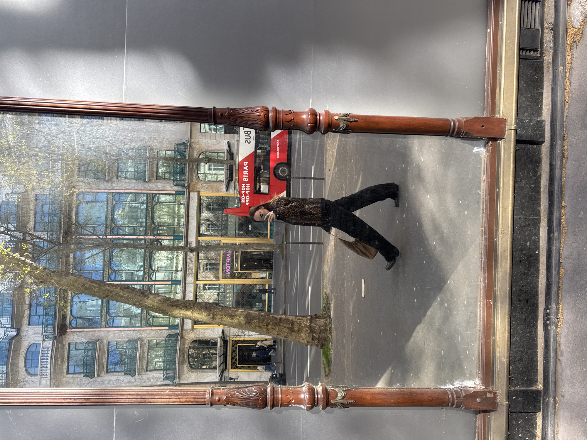The image displays a street scene in Paris, France, captured as a reflection in a large, framed mirror or window. The foreground is defined by two ornate, dark wooden vertical pillars with carved embellishments, forming the frame of the reflective surface, set upon a dark granite base with a brass-colored trim.
Reflected in the mirror, a female individual is captured in motion, walking from left to right across the lower-middle portion of the frame. She wears a long-sleeved, leopard-print top, dark wide-leg trousers, and dark flat shoes. Her right arm is raised, holding a smartphone, indicating she is taking a mirror selfie. A brown tote bag is slung over her left shoulder.
The reflected background reveals a bustling Parisian street. Across the street stands a grand, multi-story Haussmannian-style building, characterized by its ornate stone facade, numerous arched and rectangular windows, and decorative balconies. At ground level, the reversed neon sign "SPMETNIRP" (Printemps) is prominently visible, identifying the location as being near a Printemps department store. A large, deciduous tree with nascent spring foliage stands on the sidewalk, partially obscuring the building. On the asphalt street, a red double-decker tourist bus, bearing the reversed text "FFO-NOH-POH SIRAP" (Paris Hop-On Hop-Off) and "BUS 219," is positioned behind the walking individual. White lane markings are visible on the street. Several indistinct figures are present on the far sidewalk, walking in front of the Printemps building. The scene is brightly illuminated by direct sunlight, casting distinct shadows and highlighting the texture of the street and buildings.