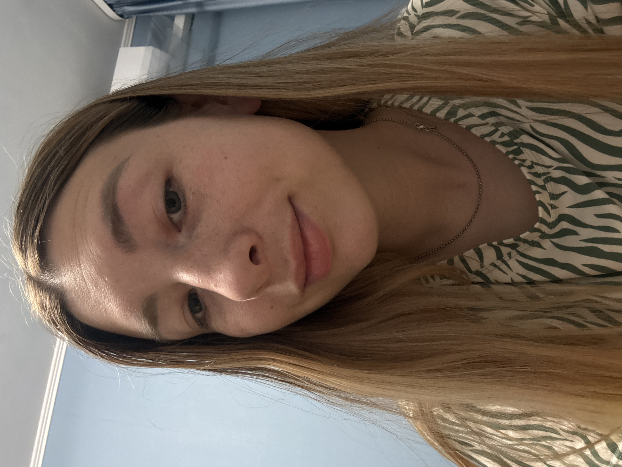 A close-up, slightly angled, indoor shot of a young woman with fair skin and light brown, straight hair. She is wearing a light-colored top with a dark green zebra stripe pattern and a thin silver necklace. Her facial features are visible including freckles, light eyebrows, and green eyes. The background includes a blue wall and window frame. The location is not identifiable as Moscow, Russia, from the given image.