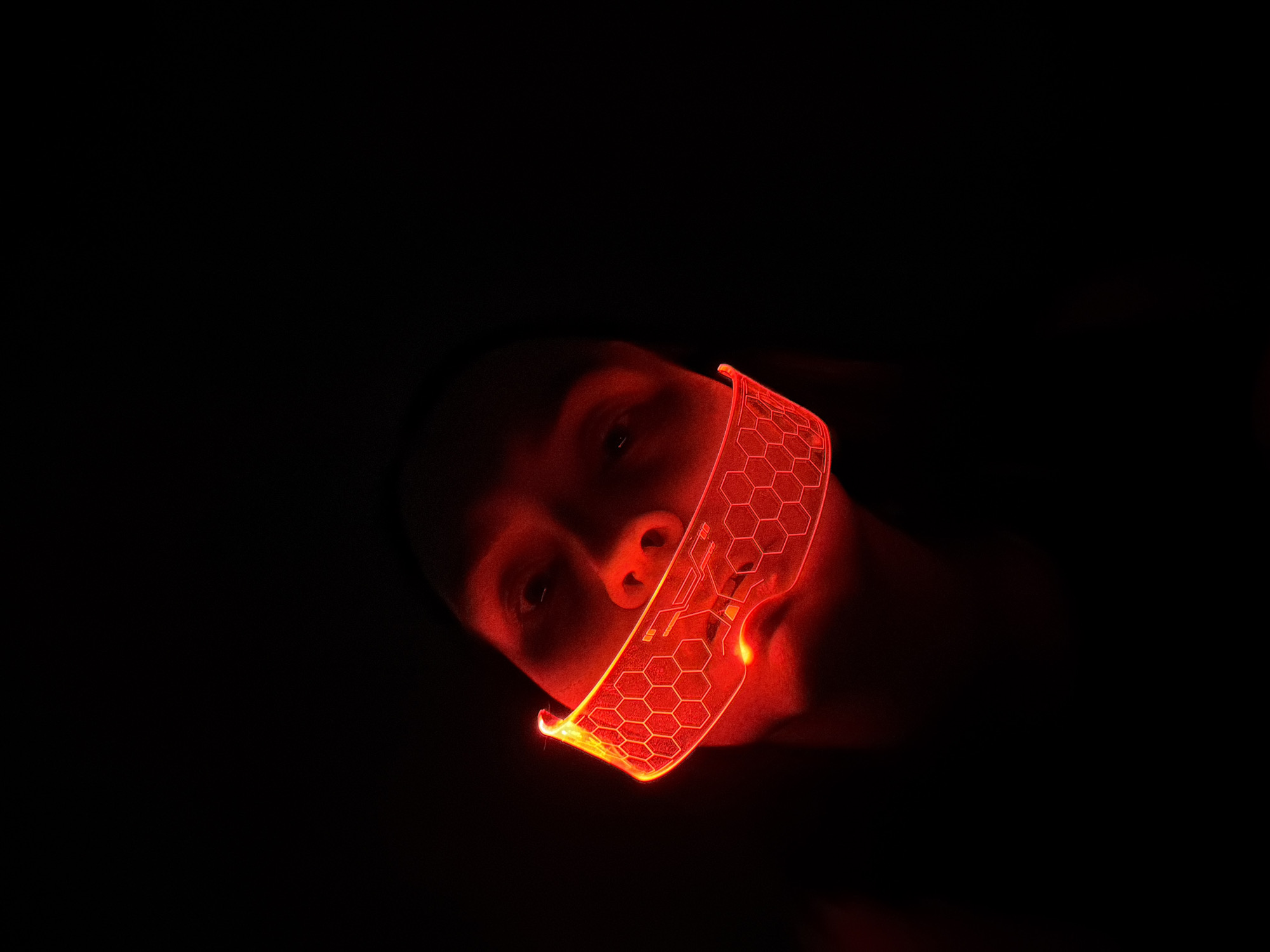 A person wearing a red, futuristic-looking mask with a hexagonal pattern lies against a dark background. The mask illuminates the person's face, revealing their nose and eyes. The scene is primarily illuminated by the red light emanating from the mask. The context of Chaikovsky, Russia is not explicitly discernible from the image.