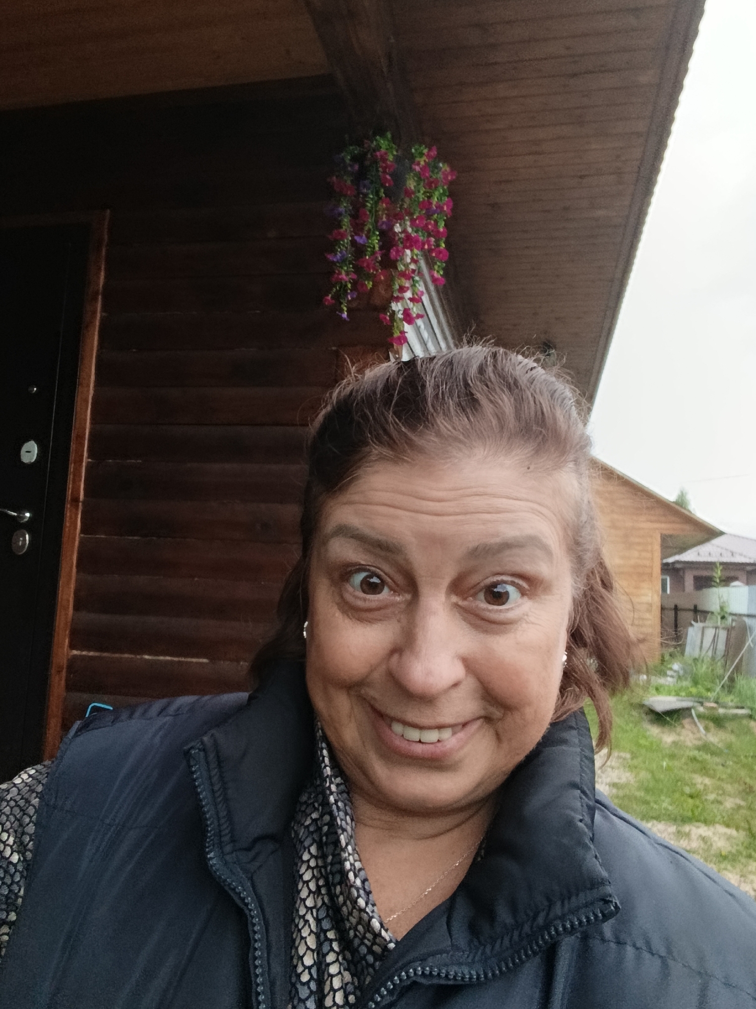 A woman is taking a selfie outdoors in front of a wooden building. She is wearing a dark vest over a patterned shirt and a necklace. She has brown hair and is smiling broadly with her eyes wide open. Above her, a hanging basket of pink and purple flowers is visible. The background shows more wooden structures and greenery. The location is in the city of Verеya, Russia.