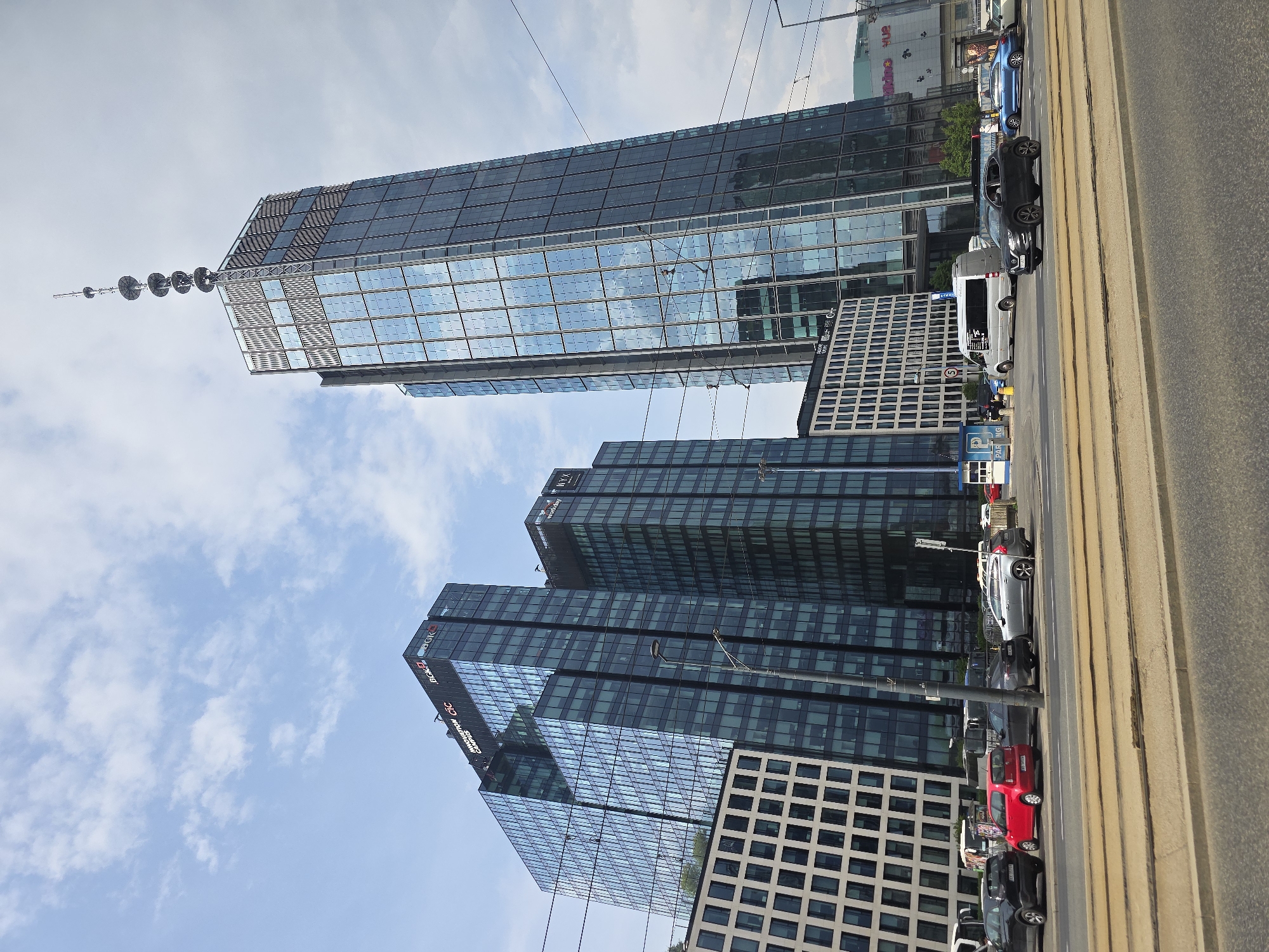 The image shows a street view in Warsaw, Poland, featuring modern architecture. The main subjects are several tall, glass-facade buildings, reflecting the sky and clouds. One building has a distinctive antenna or tower-like structure on top. Below, there is a street with vehicles, including cars and a van, parked along the side. The roadway also has tram tracks. The sidewalk is partially visible on the right side. The sky is partly cloudy.