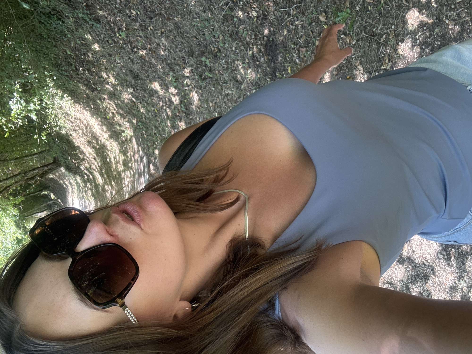 A woman with brown hair, wearing large brown sunglasses, a gray tank top, and jeans, stands on a path in a wooded area. Sunlight filters through the trees, casting shadows on the ground. She is looking down and to the left, with her lips slightly parted. Her left hand is visible in the lower left corner of the frame, and her right hand is extended to the right. The path appears to be made of dirt and gravel. The environment suggests a natural, outdoor setting. The image does not provide any specific indicators of the location being in the city of Ravilloles, France.