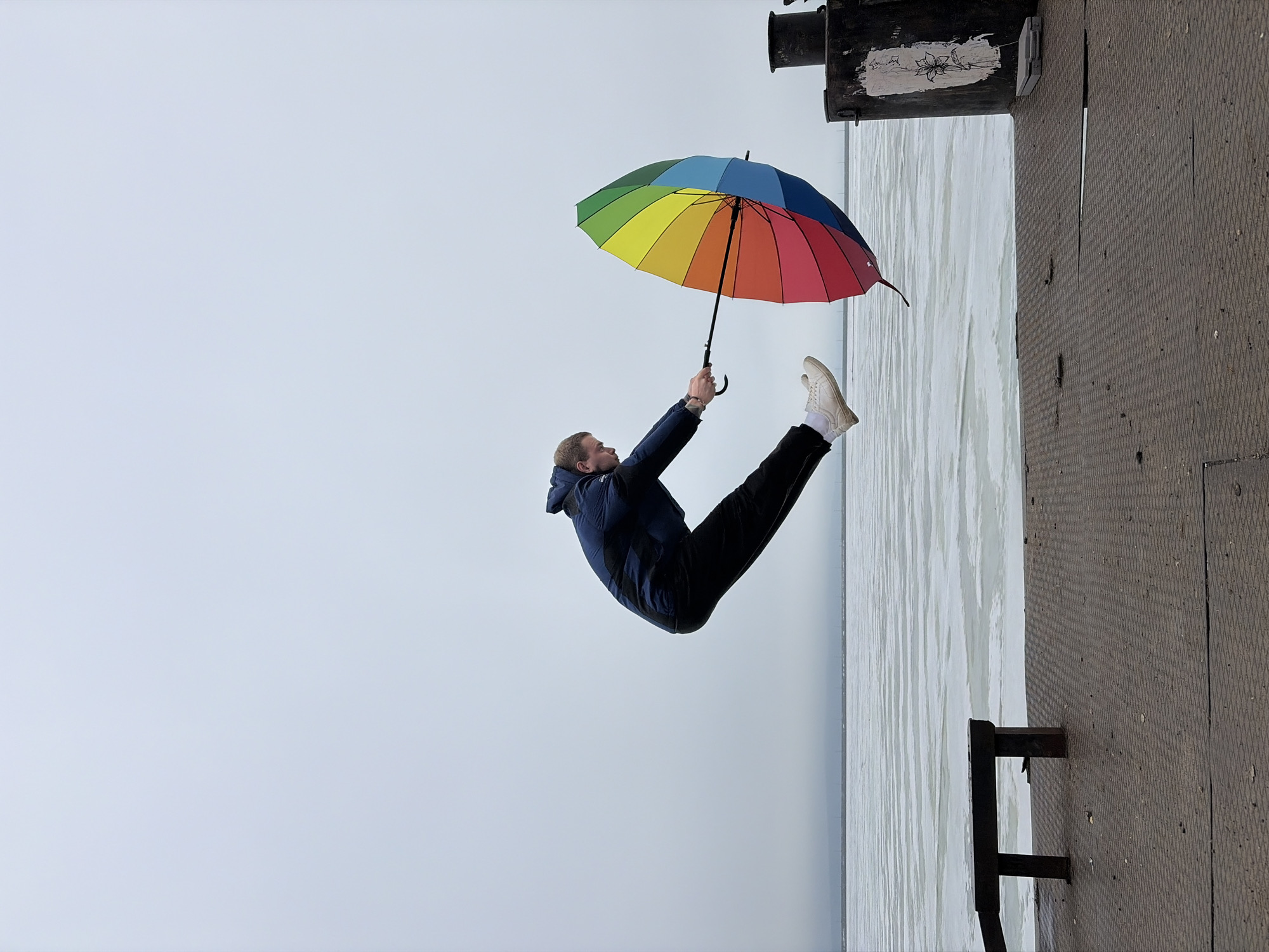 A man in a dark jacket and dark trousers with white shoes is holding a rainbow umbrella. The scene appears to be in Chaikovsky, Russia. The man is suspended in the air next to what appears to be water and a pier. The photograph is rotated 90 degrees clockwise.