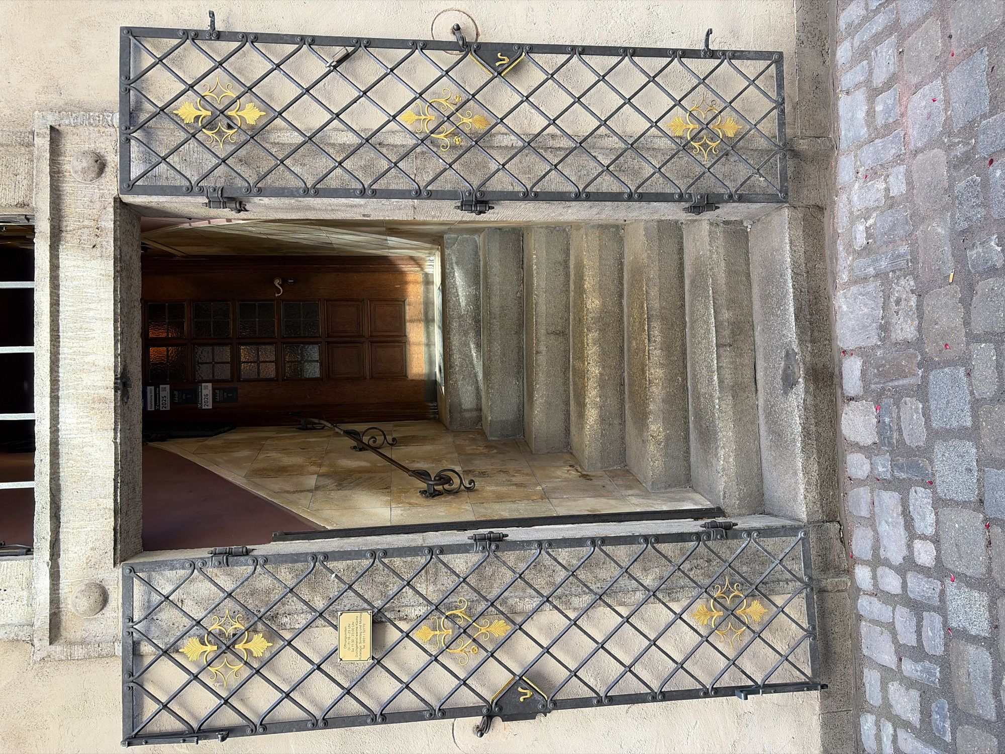 The image shows an exterior view of an entrance to a building in Würzburg, Germany. The entrance is recessed and approached by a set of stone stairs. The stairs lead up to a wooden door with glass panes. On either side of the entrance are large, ornate metal grilles with a diamond pattern, each adorned with a golden fleur-de-lis design. The grilles are open, revealing the entryway. A sign with text in German is attached to the left grille. The ground in front of the entrance is paved with cobblestones. The walls of the building are a light, textured material, and the architectural details suggest an older, possibly historic, structure. There are no people visible in the image.
