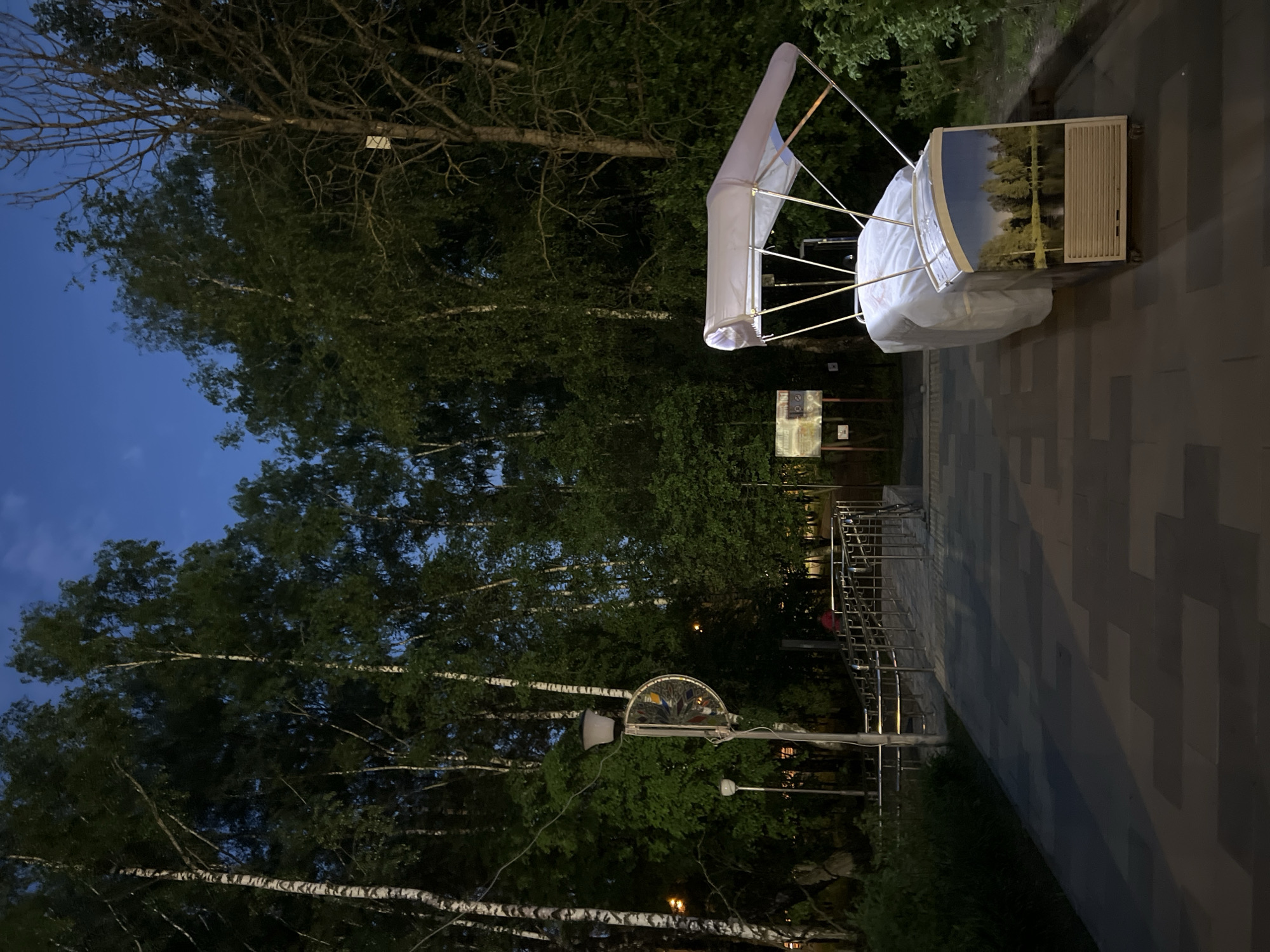 The image shows an outdoor scene in Vnukovo, Russia, at dusk or night. There is a large art installation or sculpture visible on a paved area. The installation is a complex structure made of metal and fabric, possibly illuminated from within, with a scenic background panel depicting a natural scene. In the background, there are trees with dense foliage. A bike rack made of metal bars is also visible. The ground is paved with rectangular stones of varying shades, and the lighting is dim.
