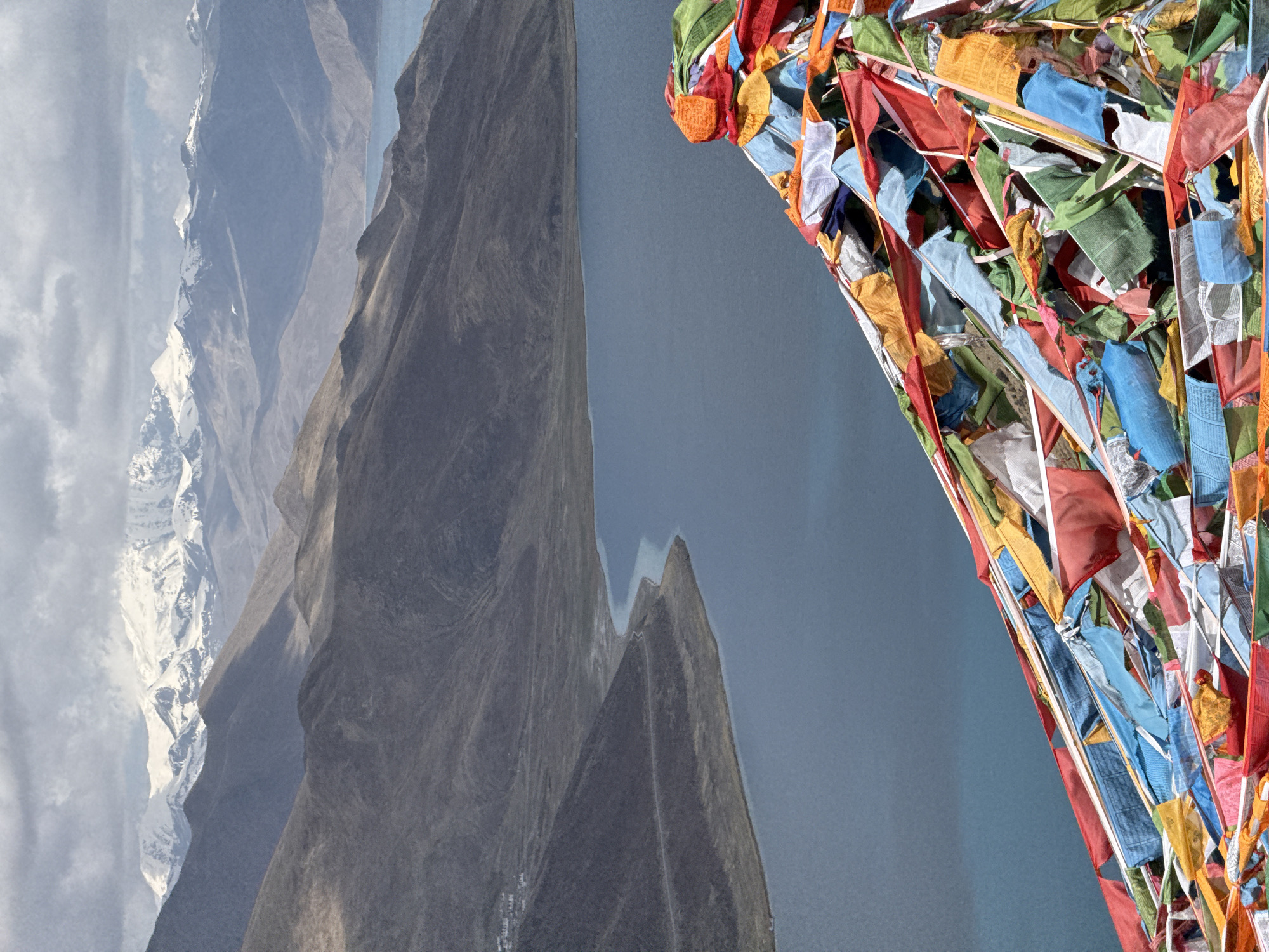 The image features a combination of natural and cultural elements, likely taken near Qangtang town, China. The left side of the image shows a mountainous landscape, with peaks of varying heights. Some of these peaks are covered in snow, while others are rocky and barren. There is a body of water, possibly a lake or river, separating the mountains from the foreground.
On the right side, there is a cluster of colorful Tibetan prayer flags strung together. The flags are multi-colored and fluttering in the wind. The background behind the flags appears to be the same body of water visible on the left side, indicating that the flags are positioned along the shoreline or a nearby elevated point.