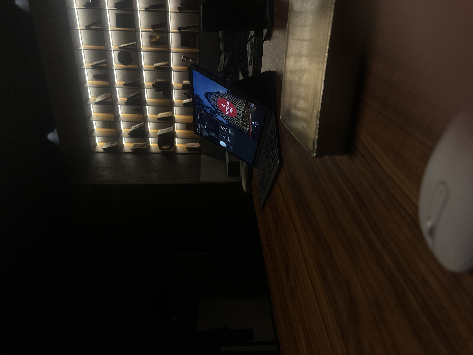 The image shows a dimly lit interior with a warm color scheme. A tablet with a keyboard is on a dark wooden table. The screen of the tablet displays a building, marked with "MICHELIN 2025." Behind the tablet, there is a shelf display filled with various artifacts, illuminated by vertical lights. To the right of the tablet, a computer mouse is placed on the table. Considering the display on the tablet and the background decor, the location appears to be the Michelin Guide Restaurant selected in the city of Sofia, Bulgaria.