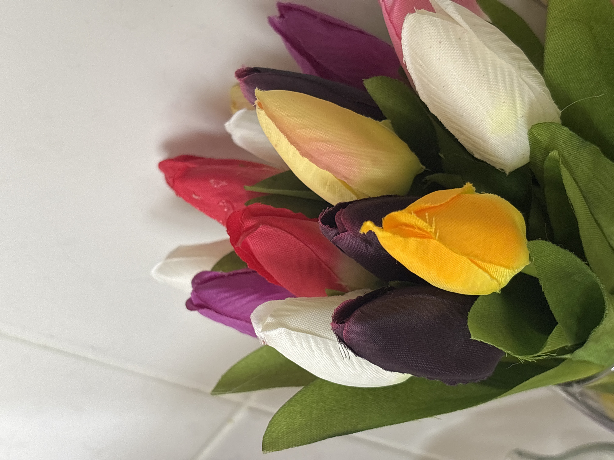 A close-up shot shows a bouquet of fabric tulips in various colors (purple, white, red, yellow, and pink) with green fabric leaves. The tulips are tightly grouped, with some partially overlapping others. The background is a plain, white surface with a slightly reflective quality. No people are visible. The location is undetermined, and the claim of Chaikovsky, Russia, is unsubstantiated.