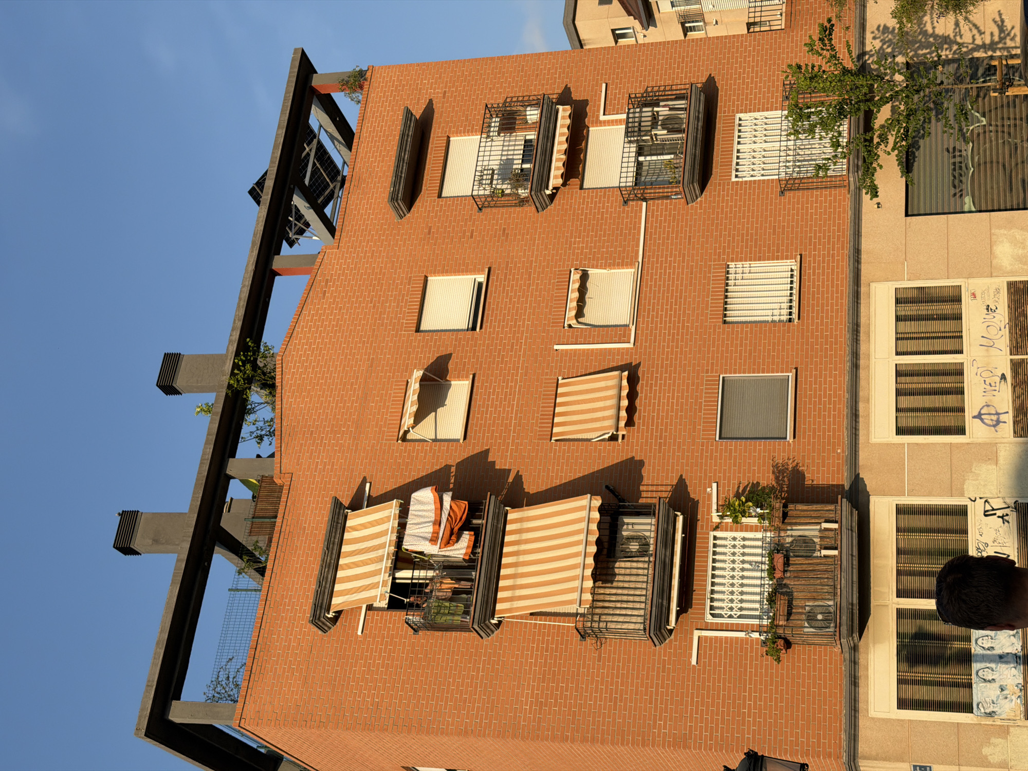 This media file captures a view of an apartment building in Benimàmet-beniferri, Spain, likely taken from a higher vantage point, possibly another building or drone. The building's facade is constructed with red bricks, and it features multiple windows, some with awnings. Several windows have metal grilles or security bars, and some are equipped with air conditioning units. There are also balconies with some greenery, suggesting residential occupancy.
The scene depicts the exterior of the building under a clear blue sky, indicating a sunny day. The sunlight casts shadows on the brickwork, suggesting it is either morning or late afternoon. The composition is slightly tilted, giving a dynamic perspective of the architecture.
On the far right of the image, a portion of another building is visible, which appears to have a lighter-colored facade and larger windows, some adorned with graffiti. A person's head is visible in the bottom right corner, looking towards the building.
One of the windows on the brick building has laundry hanging on a rack, and another has a blanket draped over it, implying everyday life activities within the apartments. The overall impression is a slice of urban residential life in Spain, characterized by functional architecture with decorative elements like awnings and security features. No discernible text is visible in the media file, aside from what appears to be graffiti on the adjacent building.