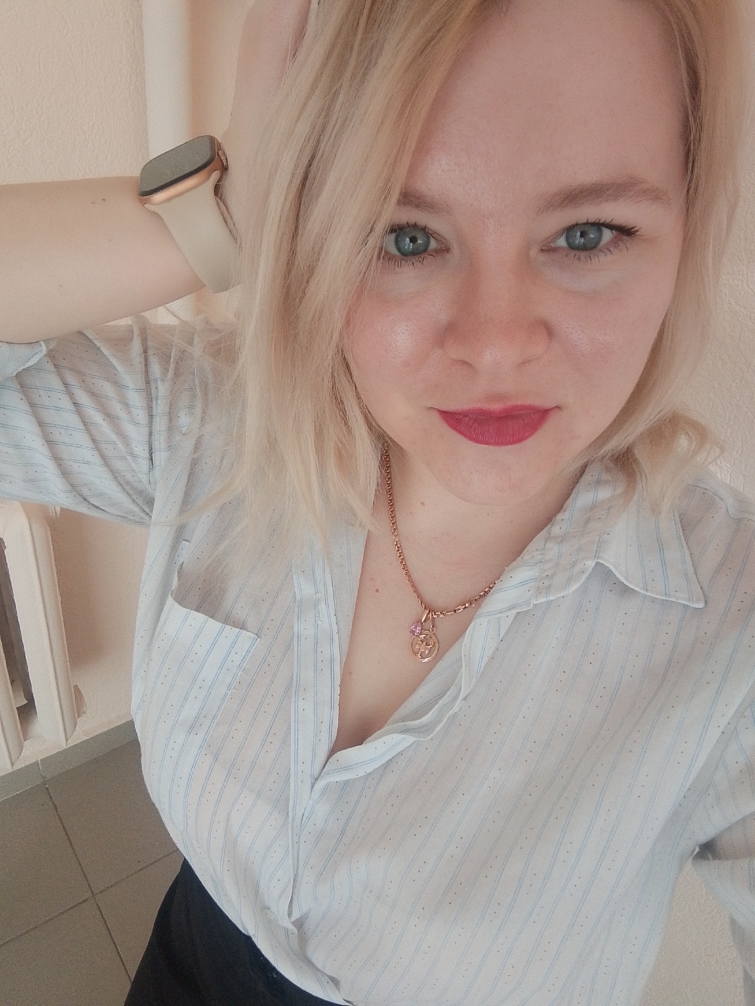 A woman with blonde hair and blue eyes is posing for a selfie. She is wearing a light blue and white striped collared shirt, unbuttoned at the top, and a gold necklace with a floral pendant. She has a gold wristwatch on her left wrist. Her lipstick is a bright pinkish-red. The background includes a wall painted light beige, a radiator, and tiled floor. There is no information to indicate that this image was taken in Izhevsk, Russia.