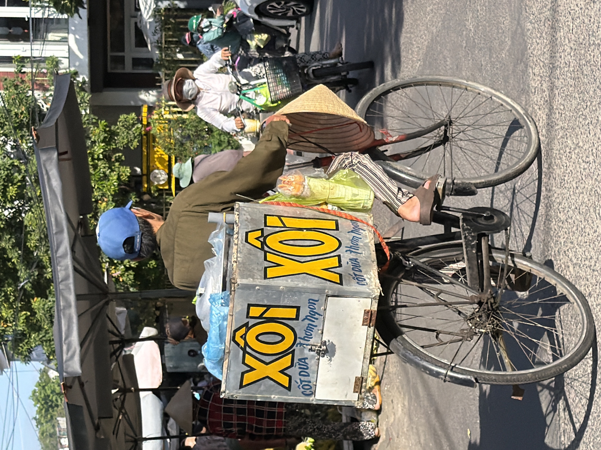 The image depicts a street scene in Nha Trang, Vietnam, where a person on a bicycle transports a large metal box on the back. The box is adorned with bright yellow lettering that reads "XÔI" with additional text in blue below. The bicyclist, seen from behind, wears a dark green long-sleeved shirt and a blue baseball cap. They are also carrying a conical hat and wearing striped pants and sandals. The background shows a market or street vendor area with stalls and awnings. Other individuals are visible, some on motorcycles, and the overall setting suggests a bustling urban environment with clear daylight.