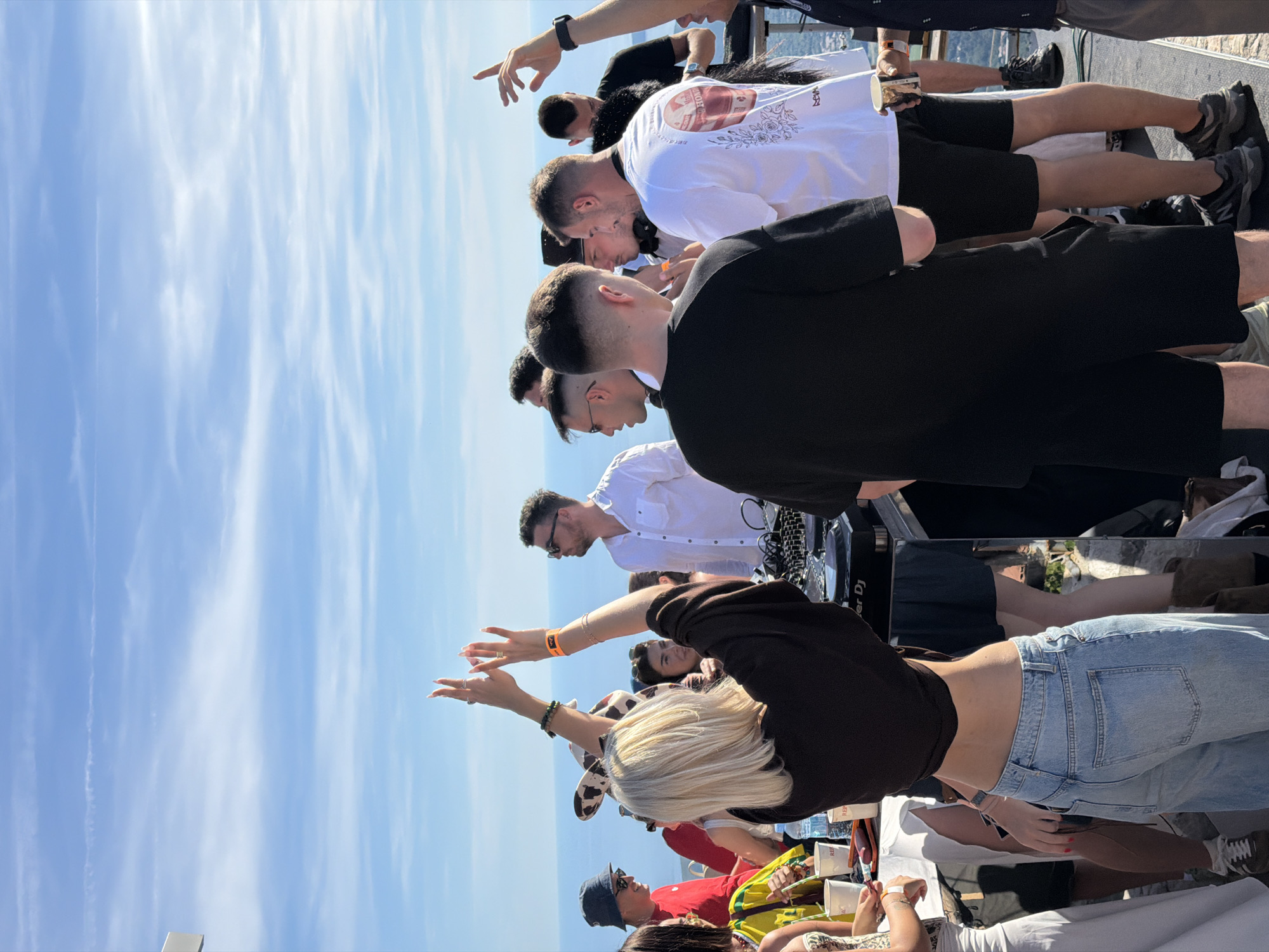 In Žanjev Do, Montenegro, on a bright, sunny day with a clear blue sky and wispy clouds, a group of people are gathered outdoors, seemingly at a party or event. The setting appears to be elevated, possibly a mountaintop or a terrace, with a view of the sea in the background.
In the foreground, a woman with blonde hair is standing with her back to the camera, wearing a dark-colored top and light-wash jeans. Her arms are raised, with her hands open, as if in a gesture of celebration or excitement. She is also wearing jewelry on her hands and wrist.
To her left, several other individuals are visible. A person wearing a red shirt and a blue bucket hat is interacting with someone in a yellow and green patterned jacket. Another person, a woman, is partially visible behind them.
To the right of the blonde woman, a DJ setup is visible, with turntables and other equipment on a table. A man in a white shirt and sunglasses is standing near the DJ setup. Further back, a group of men are standing and talking. One man in the foreground is wearing a black t-shirt, and another is wearing a white t-shirt with a graphic print.
The overall atmosphere suggests a lively outdoor gathering. The presence of the DJ equipment indicates music is being played, and the raised hands of the woman in the foreground, along with the social interactions among the people, suggest a celebratory mood.