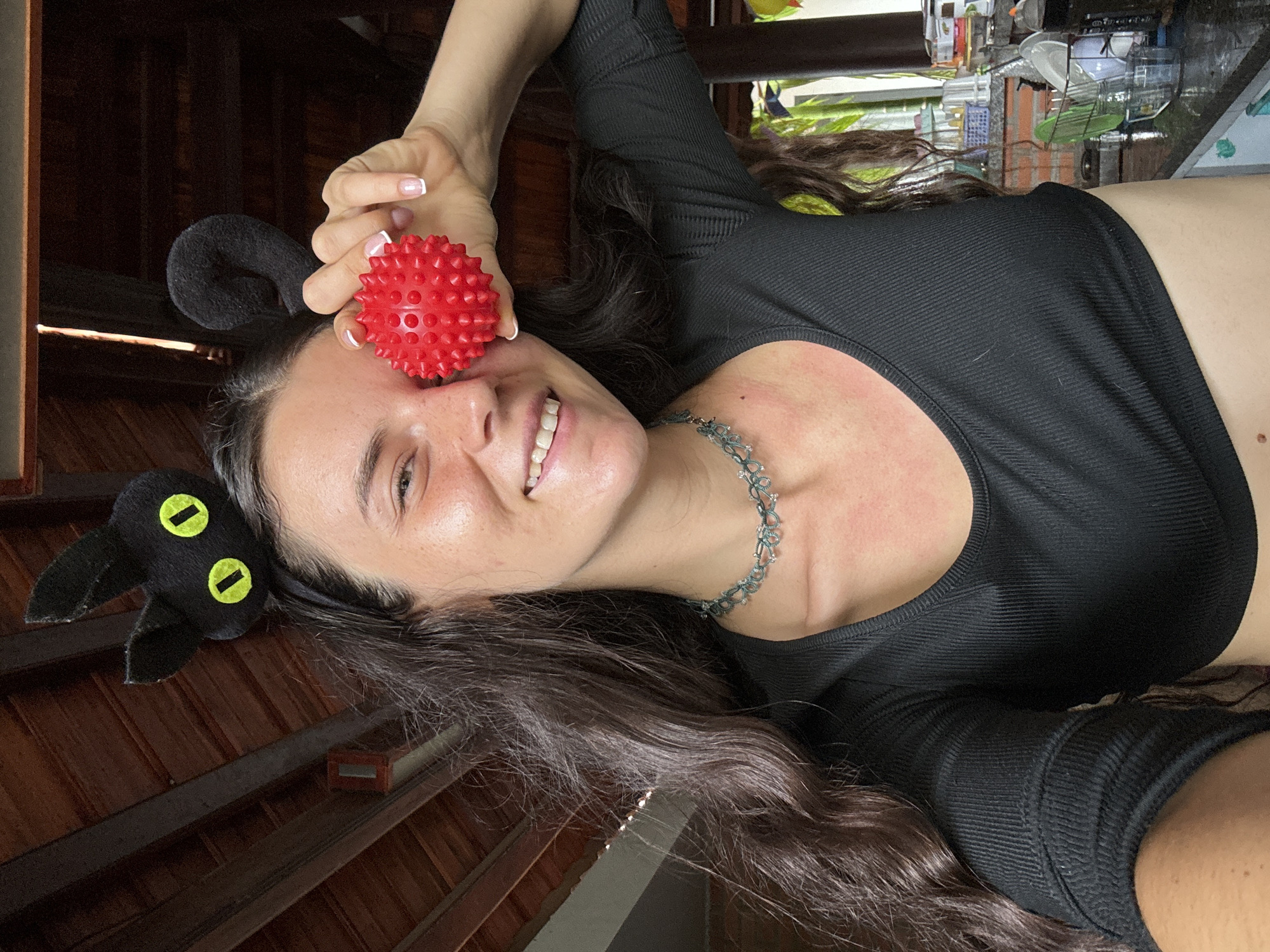 This media file presents an angled selfie portrait of a young woman, likely taken indoors or under a covered structure in Mũi Né, Vietnam, during the daytime. The woman is smiling broadly, looking directly at the camera, with her head tilted to the right. Her long, dark, wavy hair is spread around her.
She is wearing a black, long-sleeved, ribbed crop top, which reveals a portion of her midriff. Around her neck is a distinctive silver or grey necklace with an intricate, floral or lace-like design. On her head, she wears a black headband featuring cat ears, one of which is clearly visible with two round, bright yellow "eyes." She holds a small, spherical, red object covered in short, blunt spikes, possibly a massage ball, near her right eye. Her fingernails are manicured with white tips. Notable details include some redness on her neck and upper chest.
The background primarily shows dark wooden beams and a lighter wooden ceiling, suggesting a rustic or tropical architectural style. In the upper right quadrant, some blurred household items, possibly kitchenware like glasses and dishes on shelves, are visible. The natural lighting is bright, creating a cheerful atmosphere. No visible text is present in the image.