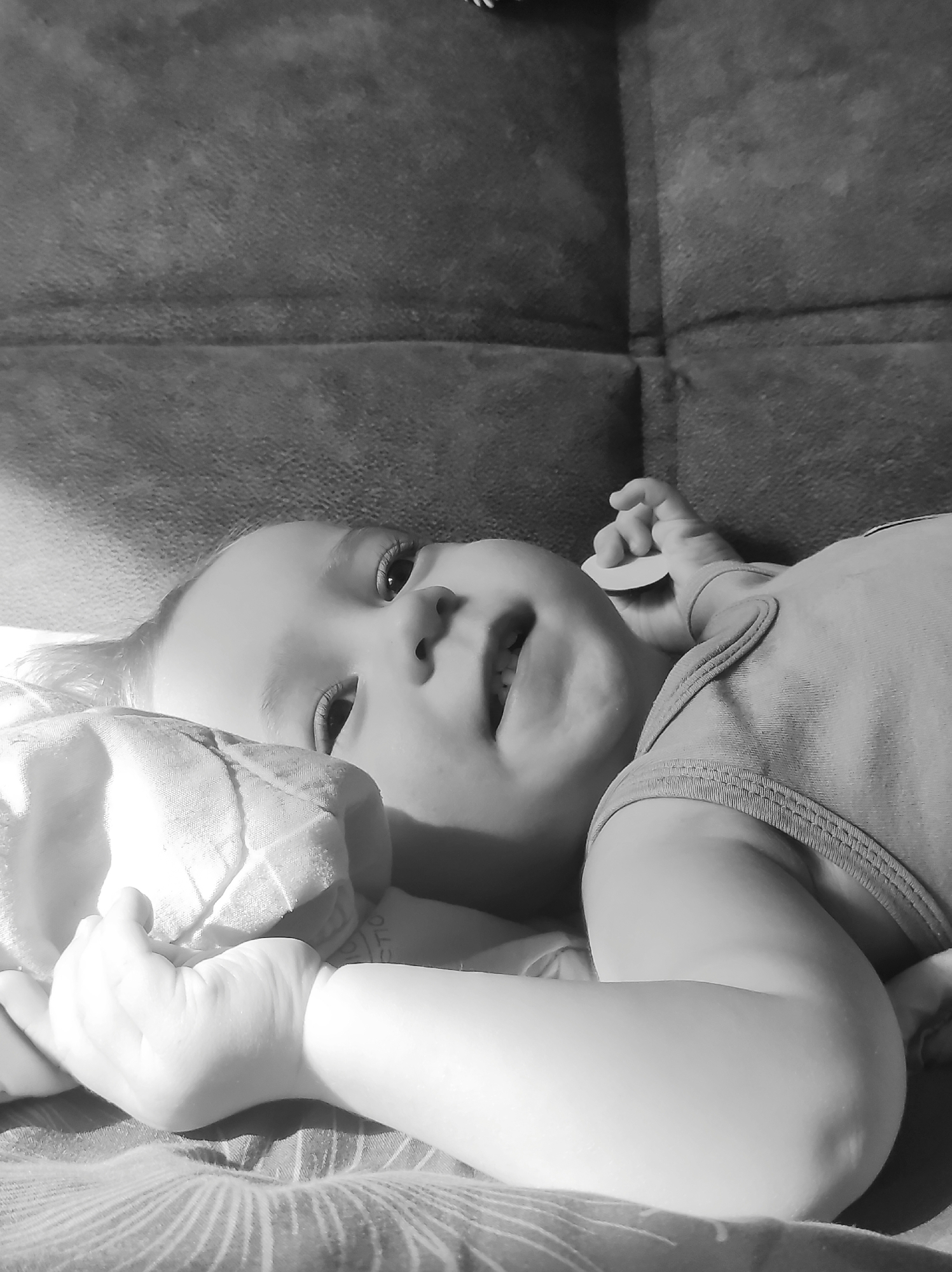 A black and white, eye-level image depicts a baby lying on their back, looking upwards and slightly to the right. The baby has wide, attentive eyes and a slightly open mouth, conveying curiosity or a mild expression. Light-colored hair is visible around their face.
The baby is wearing a sleeveless bodysuit and is positioned on a textured surface, likely a couch or bed. In their left hand, the baby holds a soft, light-colored, possibly quilted blanket or cloth. Near the baby's right hand, a small, light-colored, disc-shaped object is visible.
Strong, directional light, presumably natural sunlight, illuminates the scene from the left, creating bright highlights on the baby's skin, hands, and the soft object they are holding. This lighting also enhances the visible textures of the fabrics. The background features a darker, textured surface, possibly the backrest of a piece of furniture, while the surface beneath the baby shows faint patterned lines.
Partial text, appearing to be "CTION", is visible on a tag or label located near the baby's left hand, partially obscured by the soft cloth. The scene portrays a calm, daytime moment indoors in Chaikovsky, Russia.
