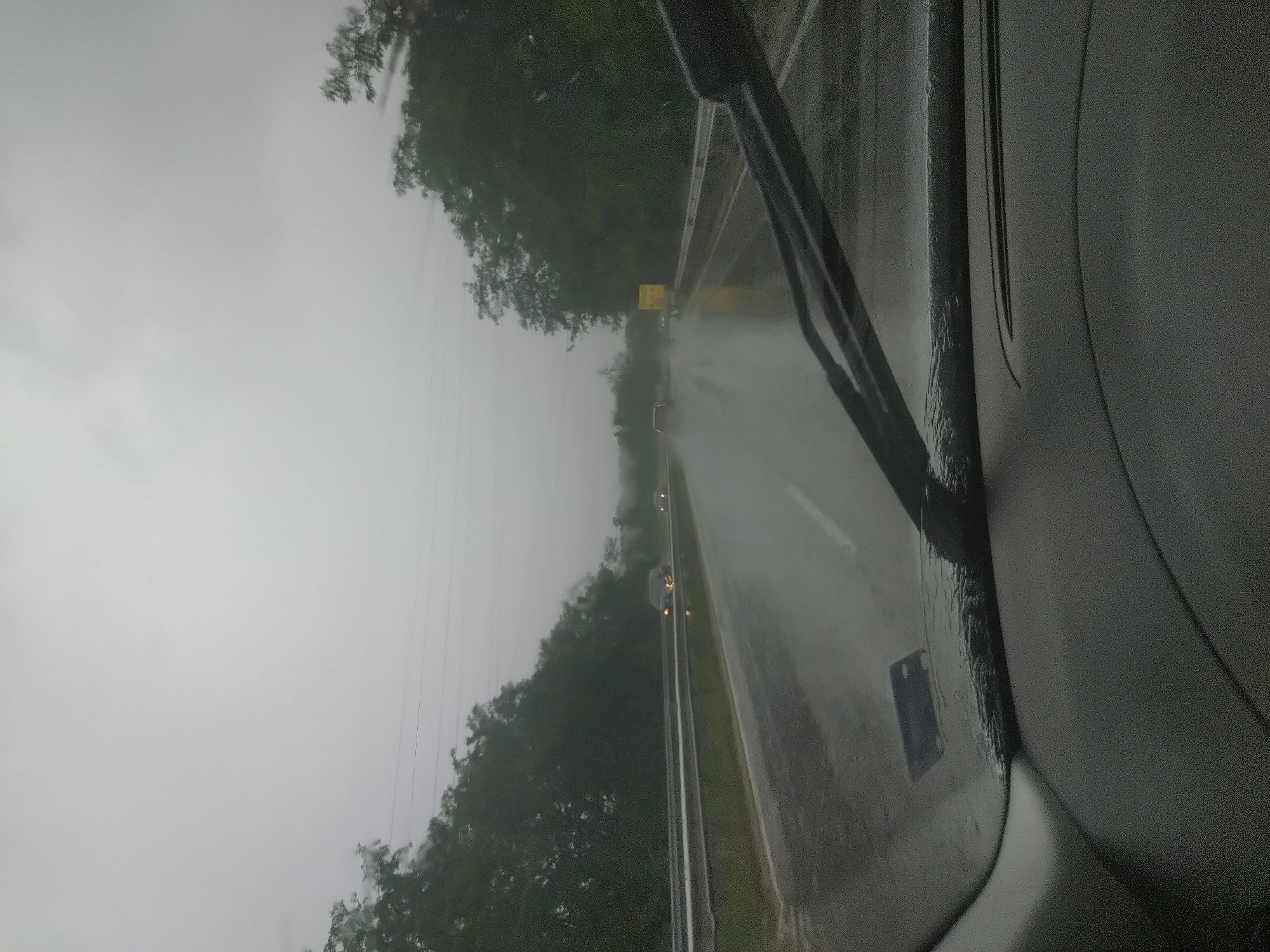 This image depicts a view from inside a vehicle traveling on a wet road during a rainstorm. The road is a multi-lane highway with a guardrail on the left side and trees lining the right side. Several cars are visible on the road, some with their headlights on. The sky is overcast and grey, indicating poor weather conditions. The foreground shows the interior of the car, including the dashboard and windshield wipers. There are no people visible. The image appears to have been taken in the vicinity of Zmeyovo, Bulgaria, as suggested by the context.