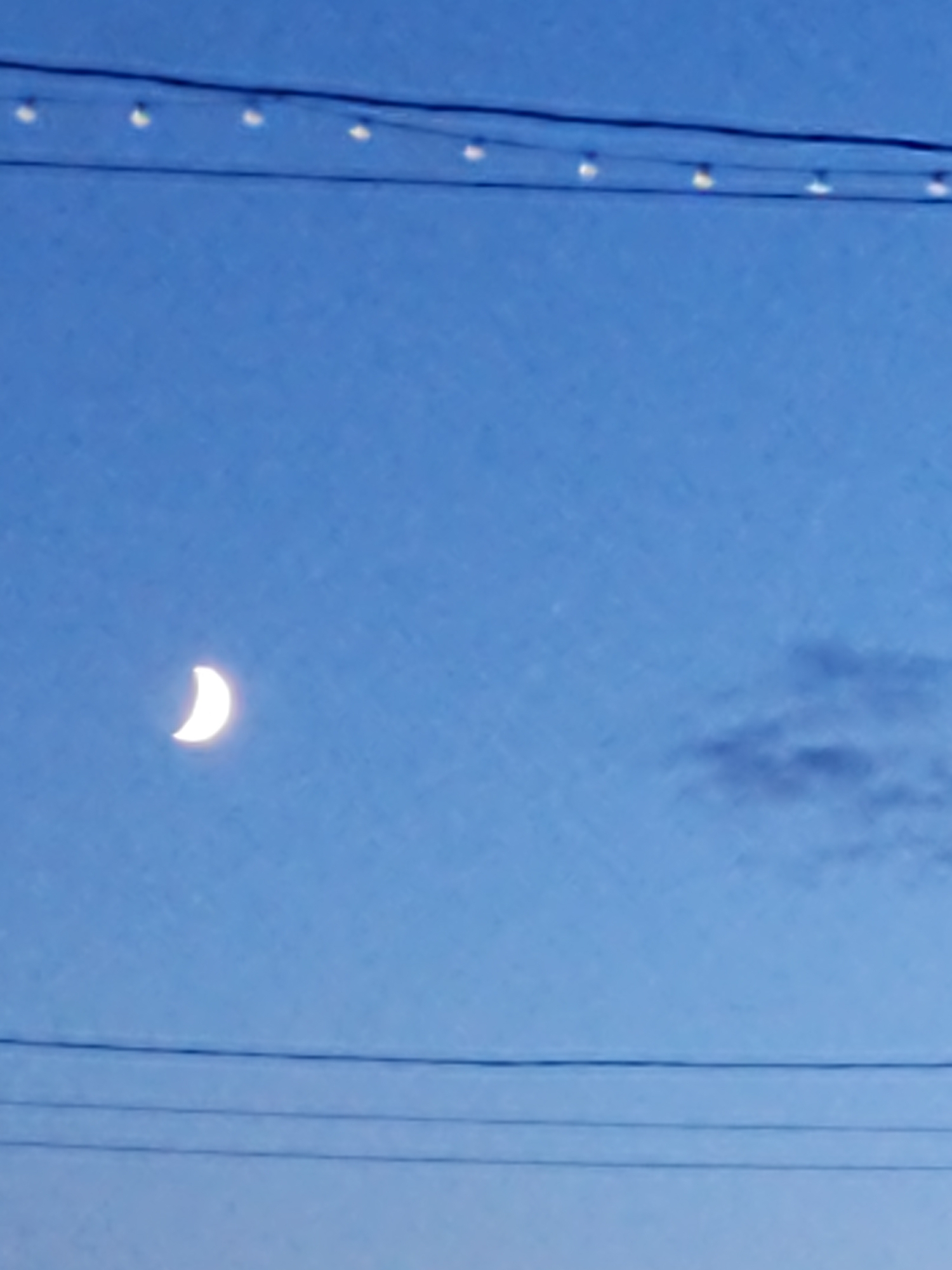 A crescent moon is visible in the clear blue sky. In the upper portion of the image, a string of lights is strung across the sky, supported by what appear to be power lines. More power lines are visible in the lower portion of the image. A faint, wispy cloud is present in the upper right quadrant of the image.