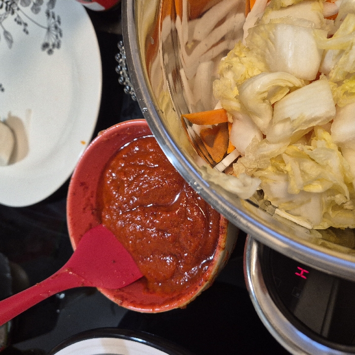 The image captures a close-up, overhead view of a food preparation scene, likely in a kitchen. The main focus is on ingredients for what appears to be kimchi or a similar fermented vegetable dish.
On the right side of the frame, a large, reflective silver metal mixing bowl is partially visible. Inside this bowl are chopped vegetables: predominantly light green and white pieces of what looks like Nappa cabbage, mixed with bright orange julienned carrots and some thinner white strips, possibly daikon radish or more cabbage stems. The bowl is resting on a black digital kitchen scale, where a red "H" is visible on its display, suggesting it's either in a "hold" state or displaying part of a reading.
To the left and slightly below the metal bowl, a vibrant red ceramic bowl is placed. This bowl contains a thick, reddish-orange paste, which appears to be a spicy seasoning mixture, likely a kimchi paste (gochujang and gochugaru-based). A bright red silicone spatula is partially submerged in this paste, with its handle extending towards the bottom left of the image.
In the upper left corner, a portion of a white plate with a delicate black floral or leafy pattern is visible. The background surface is dark and reflective, possibly a black countertop or stovetop.
The scene suggests an ongoing culinary activity, specifically the assembly of ingredients for a recipe, with the weighing of vegetables and the prepared seasoning paste ready to be mixed. The bright colors of the food contrast sharply with the dark background, highlighting the freshness of the ingredients.