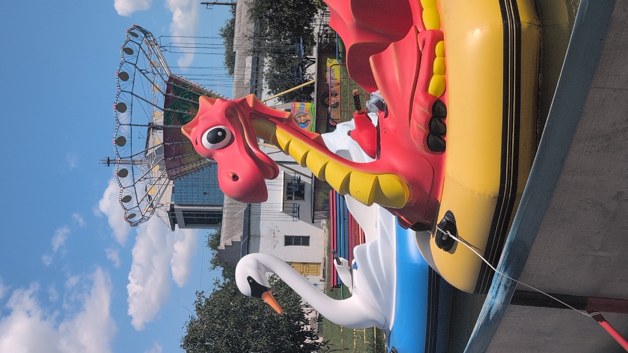 In Izmail, Ukraine, a pair of inflatable boats shaped like a swan and a dragon are docked on a body of water. The swan boat, white with a blue hull, is in the foreground on the left. To its right is a red and yellow dragon boat. In the background, a swing ride attraction at an amusement park is visible, with a building and trees surrounding it. The sky above is blue with scattered clouds.