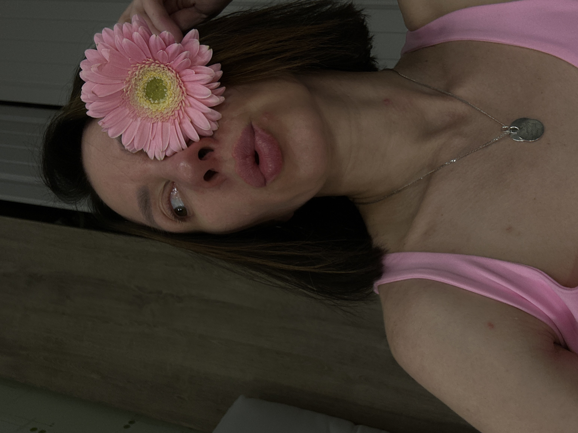 A woman wearing a pink top and a silver necklace with circular pendants poses with a pink gerbera daisy covering her right eye. Her head is tilted, and she is looking slightly to her left. She is inside, with what appears to be a wooden surface visible in the background. The location of the scene is not identifiable as Votkinsk, Russia, based on the visual content.