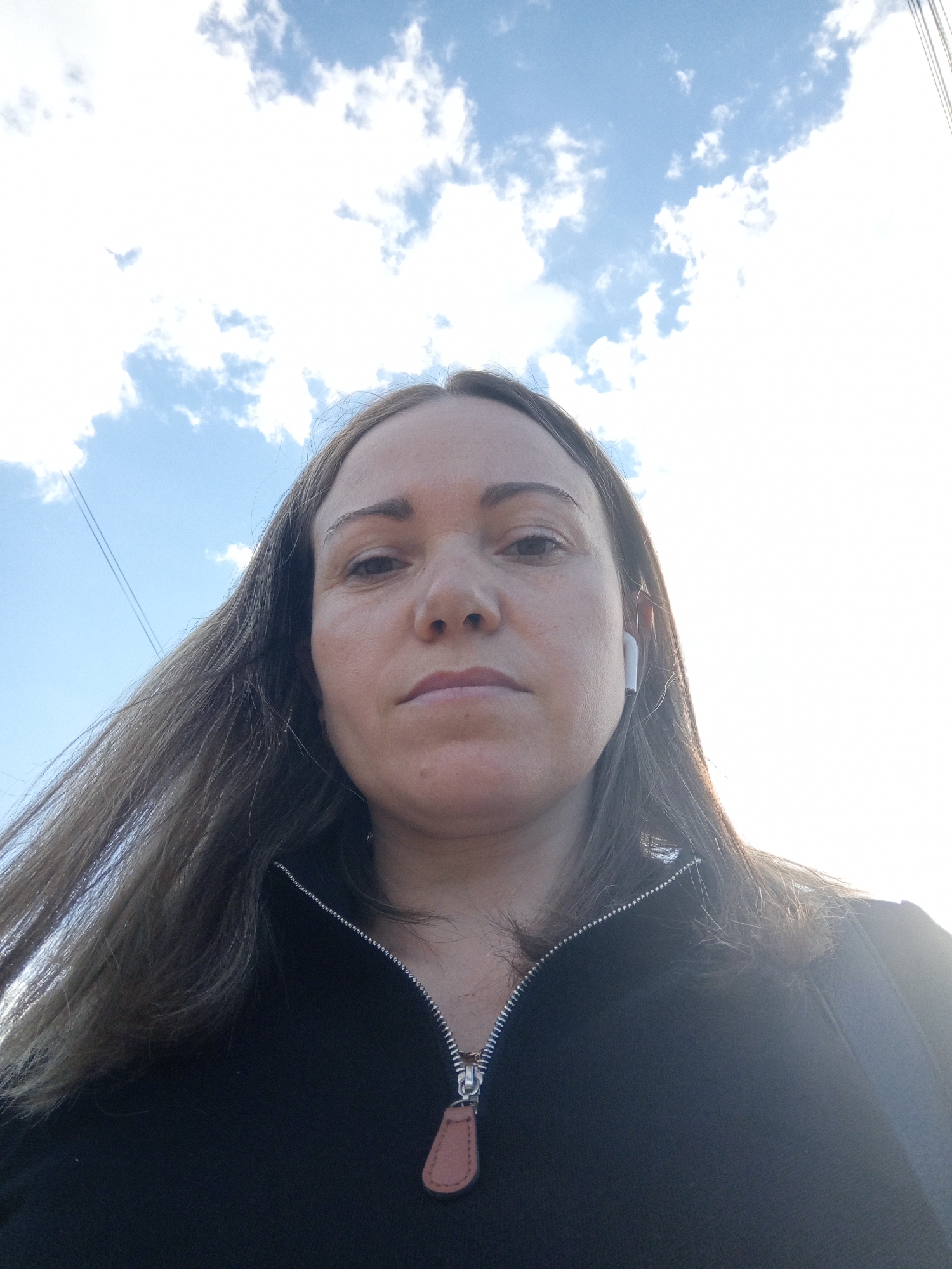 A woman, with long brown hair, is looking up at the camera from a low angle. She is wearing a black zippered top and white earbuds. The background is a bright sky with some clouds. There are power lines visible in the upper left corner. The image appears to be a selfie taken outdoors.