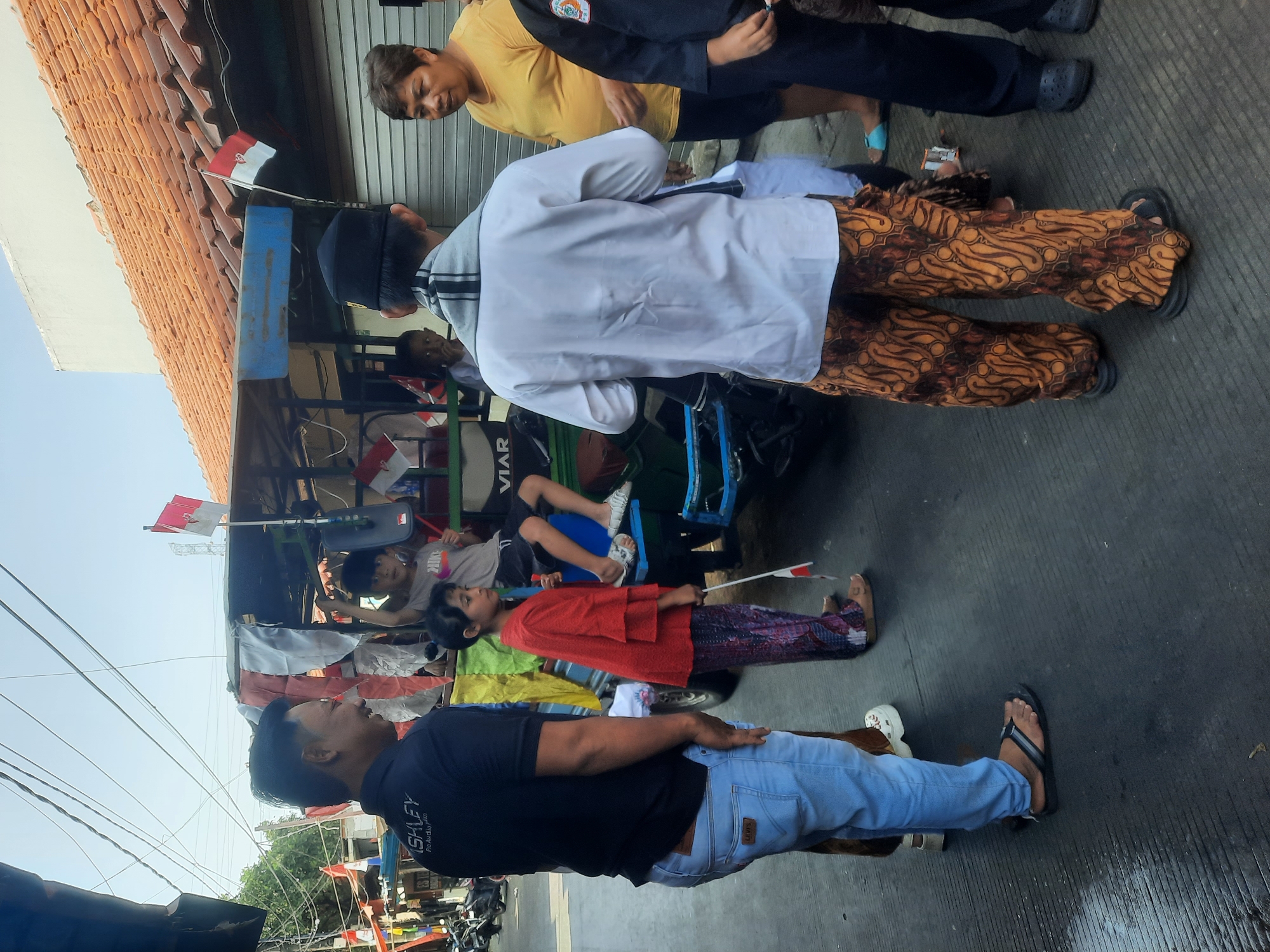 In North Jakarta, Indonesia, a group of people gathers around a small, open-air vehicle decorated with Indonesian flags. The vehicle, possibly a modified tricycle or small truck, has a canopy and seating areas. On the right, a woman in a yellow shirt looks towards the left. A man in a white shirt, black cap, and patterned pants stands with his back to the camera, facing the vehicle. To his left, a young girl in a red top and patterned pants holds a small flag. In the center of the vehicle, a boy in a grey t-shirt sits with his legs crossed, holding a flag. Another man in a black shirt and light blue jeans stands to the far left, looking towards the center. The scene takes place on a paved street, with buildings and overhead wires visible in the background. The overall atmosphere appears to be a casual gathering, possibly related to a local celebration or event, given the prominent display of national flags.