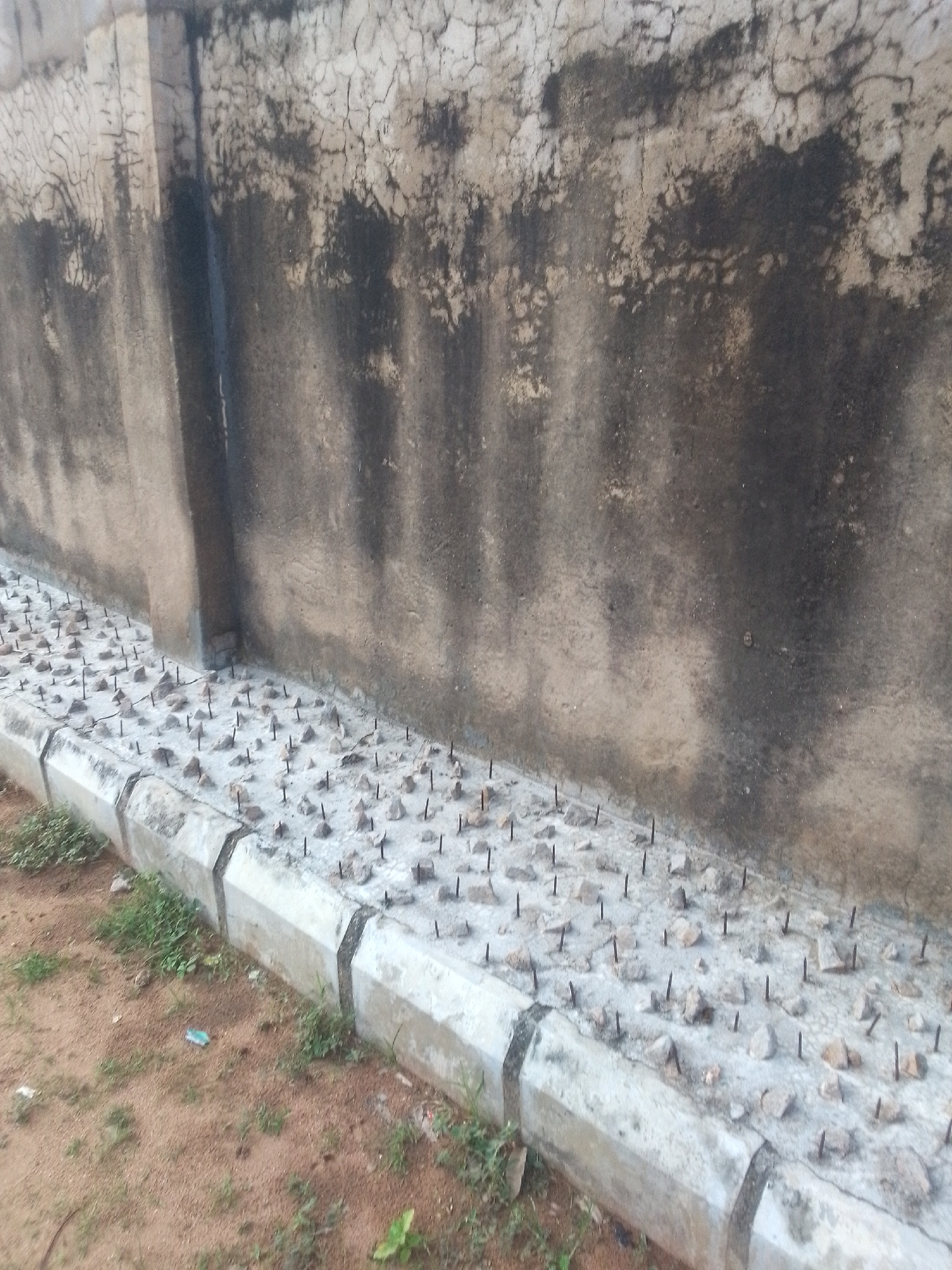 The image shows a section of a weathered, textured wall casting a shadow on a concrete border lined with numerous sharp metal spikes embedded in gravel. The wall, possibly made of concrete or stucco, exhibits a rough, cracked surface with dark stains and lighter patches, suggesting age and exposure to the elements. The concrete border is segmented with angled joints and has a slightly worn appearance. The ground around the border is a mix of sandy soil, scattered small stones, and patches of sparse green vegetation.
The spikes are arranged in a dense, uniform pattern across the gravel bed, pointing upwards, creating a deterrent. The overall impression is one of a security measure, likely intended to prevent unauthorized access or passage. The scene appears to be outdoors, possibly in a public or private space that requires protection. The lighting suggests it is daytime, with shadows indicating the direction of the sun. There are no people or animals visible in the frame. The environment seems somewhat dry, with the sandy soil and limited vegetation typical of certain climates. The focus is on the wall and the spiked deterrent at its base, emphasizing the protective or prohibitive nature of the installation. The location context of Jalingo, Nigeria, might suggest a climate or architectural style relevant to that region. There is no visible text in the image.