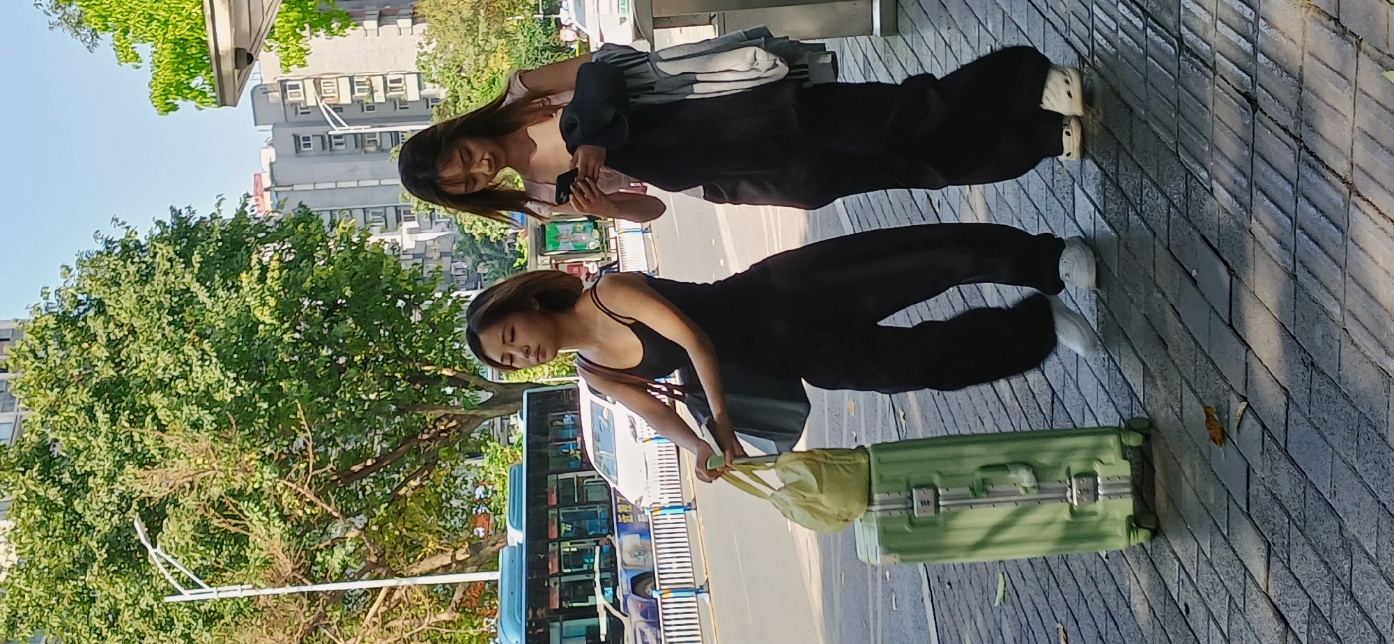 The image, oriented horizontally, captures two young women on a paved sidewalk in an urban environment, likely in Yuzhong District, China, under bright, clear daylight.
In the foreground, a woman with dark hair, wearing a black tank top and black pants, stands next to a light green hard-shell suitcase. She is holding a light yellow tote bag, suggesting she is either traveling or has recently arrived. Her gaze is directed downwards or straight ahead, and she wears white sneakers.
Behind her and slightly to the right, another woman with long dark hair, dressed in a black jacket over a lighter top and black pants, is engrossed in her smartphone. She also wears light-colored sneakers. Both women appear casually dressed in dark attire.
The background reveals a bustling city scene. Multi-story buildings with numerous windows line the street, characteristic of urban architecture. Verdant green trees with dense foliage are interspersed along the sidewalk and street, providing natural relief. A bus with blue and white accents is visible on the road behind the women, indicating active traffic. The sidewalk itself is composed of grey paver stones laid in a herringbone pattern. The strong sunlight casts distinct shadows, highlighting the clear weather conditions. There is no clearly visible text on any signs or objects. The image depicts a candid moment of two individuals navigating a sunny city street.