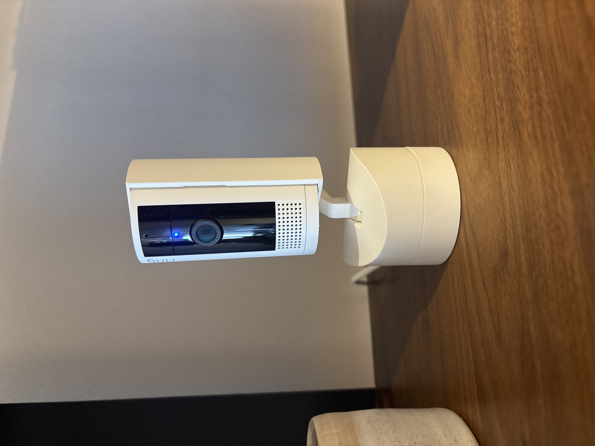 A white Ring security camera with a blue indicator light is positioned on a wooden shelf. The camera is mounted on a white adjustable stand, which is attached to a white circular base. To the left of the camera, a portion of a textured ceramic vase is visible. The background consists of a plain, neutral-colored wall. The scene appears to be indoors.